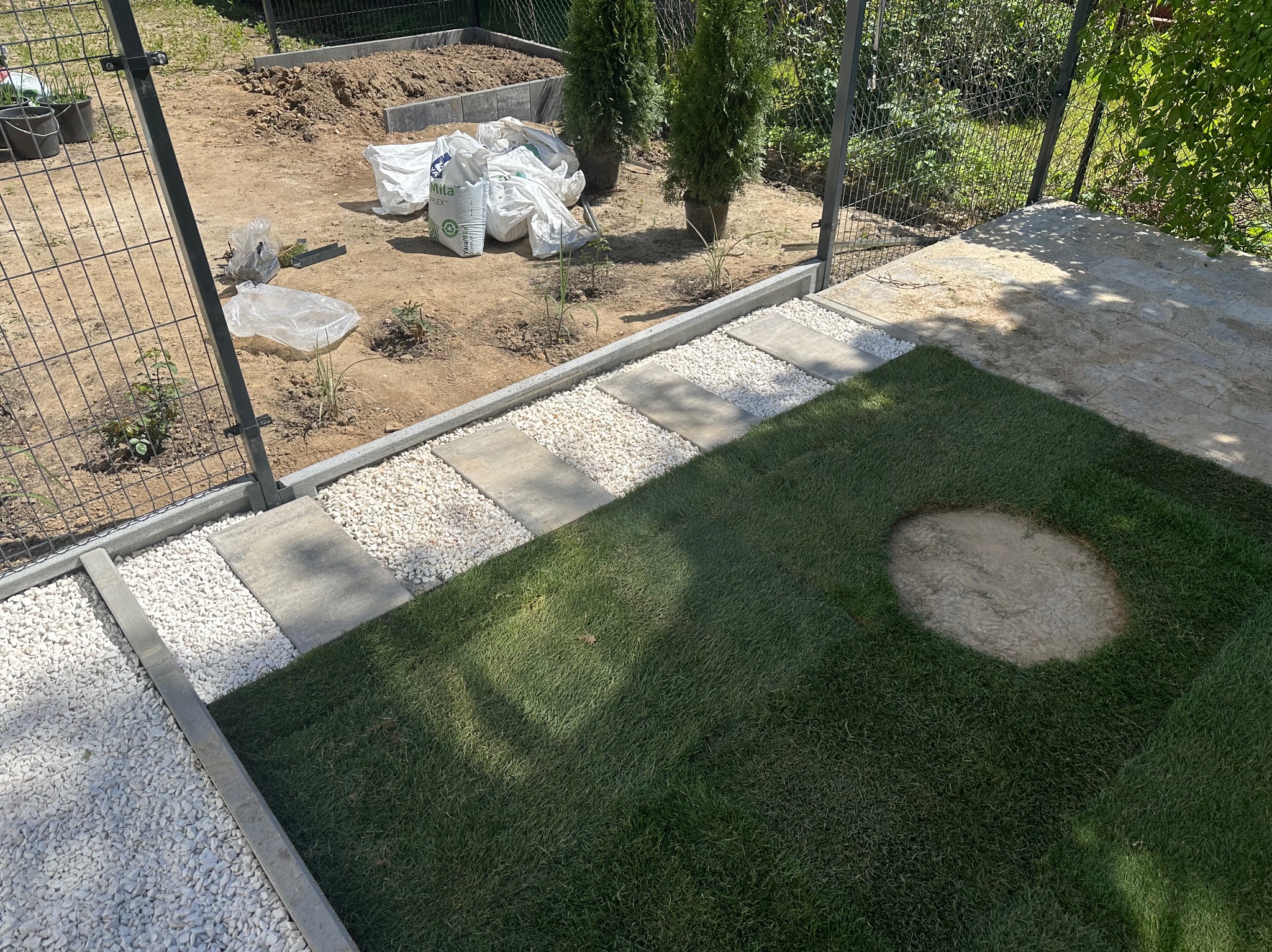 Aranżacja ogrodu z trawnikiem, ścieżką z kamieni i białego żwiru. W tle widoczne worki z ziemią i sadzonki drzew, sugerujące etap prac ogrodniczych.