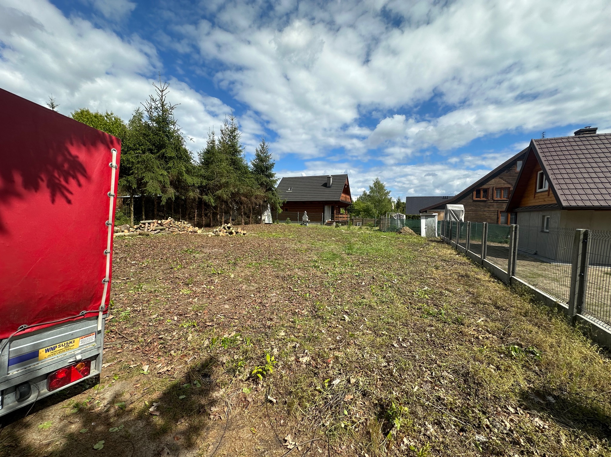 Przygotowany teren pod trawnik z widocznym stosem drewna, drzewami iglastymi i fragmentem czerwonej plandeki przyczepy. W tle domy jednorodzinne.