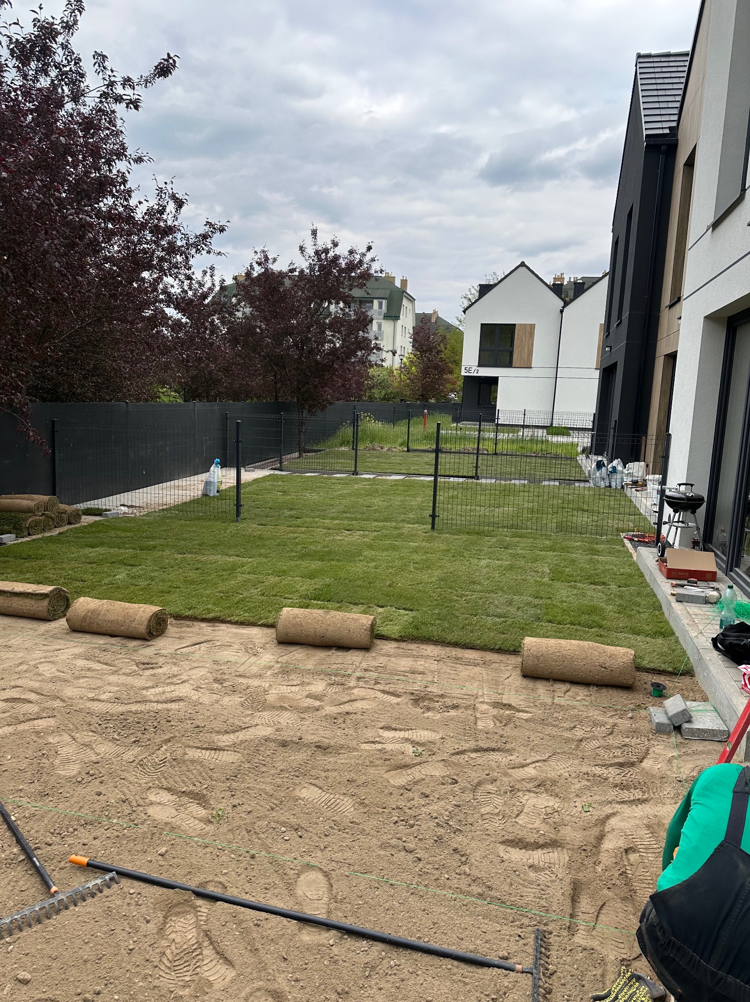 Układanie trawy z rolki w ogrodzie nowoczesnego domu. Częściowo rozłożona darń, widoczne narzędzia i rolki trawy. Czarny płot w tle, pochmurne niebo.