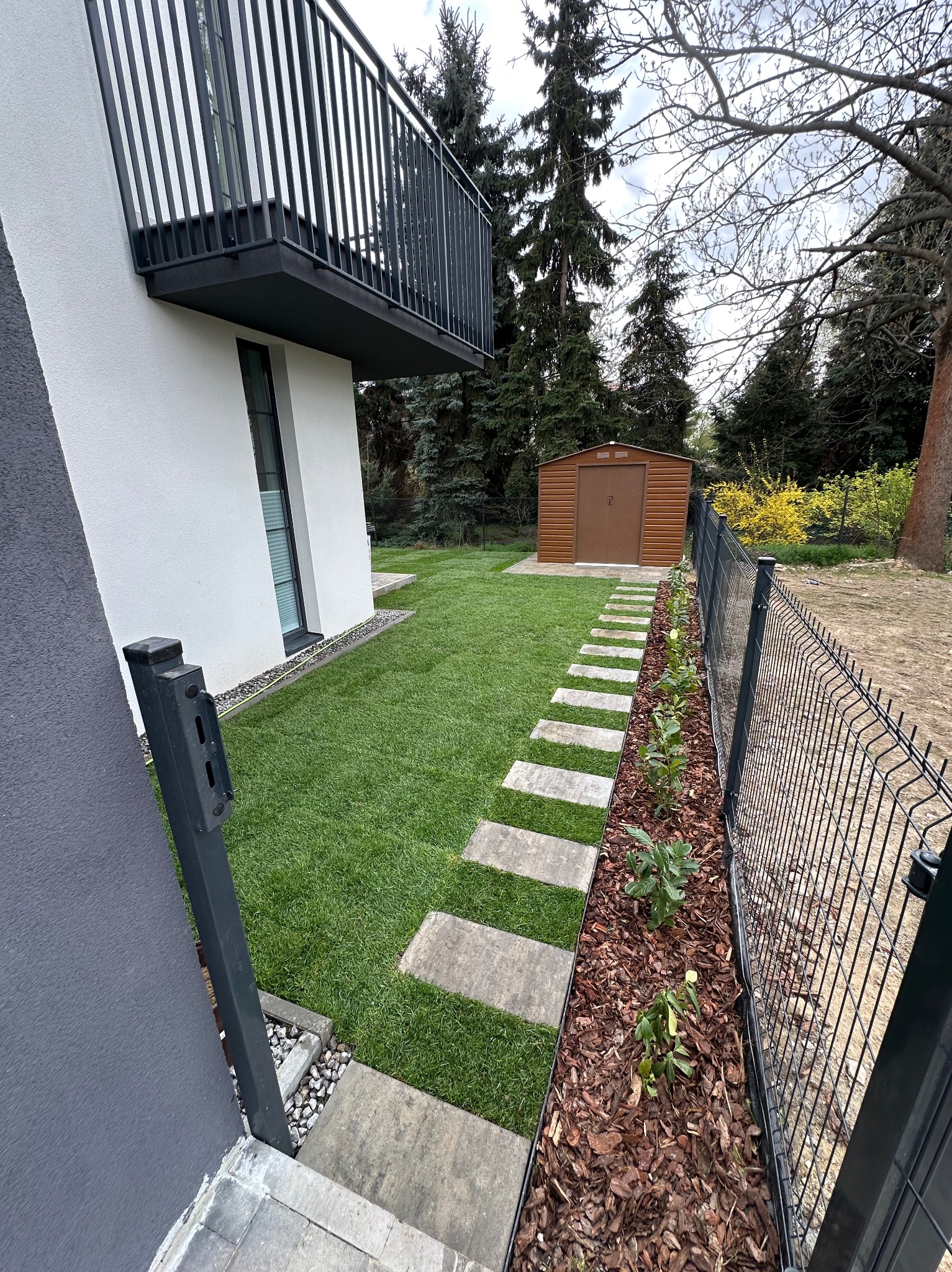 Nowoczesny ogród: trawnik, ścieżka z płyt, rabata z korą i roślinami wzdłuż ogrodzenia, brązowy domek narzędziowy w tle, fragment budynku z balkonem.