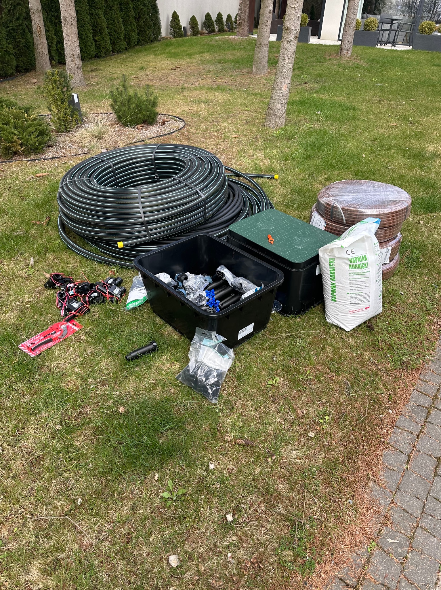 Elementy systemu nawadniania rozłożone na trawie: rura w zwoju, złączki, skrzynka sterująca i nawóz. Przygotowanie do instalacji systemu nawadniania ogrodu.