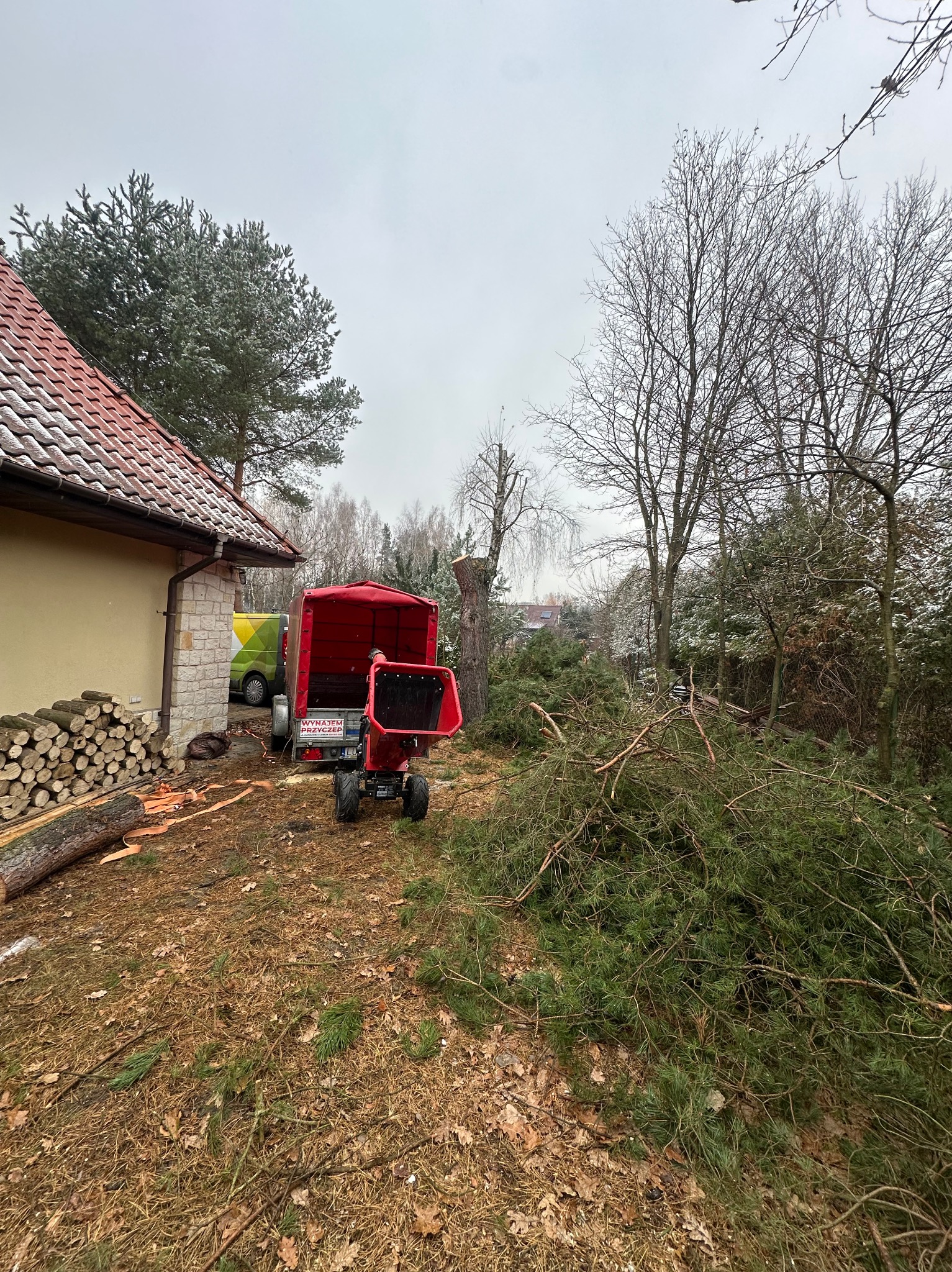 Czerwony rębak do gałęzi załadowany przyczepką, stosy pociętych gałęzi po wycince drzew na posesji z domem i stosem drewna. Praca w toku.