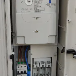 Otwarta szafka elektryczna z zamontowanym licznikiem NORAX 3 APATOR, bezpiecznikami i okablowaniem, widoczne ślady montażu.