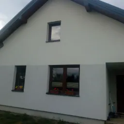 Ocieplony dom jednorodzinny z szarą elewacją, brązową stolarką okienną i kwiatami w donicach pod oknami, widok z boku.