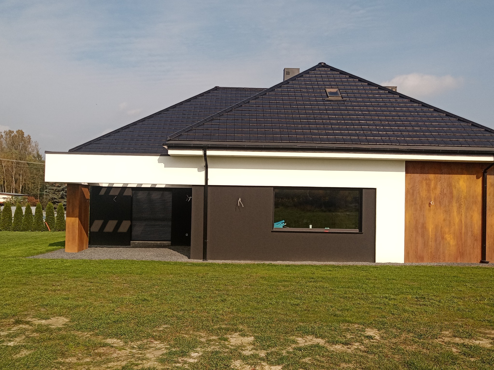 Nowoczesny dom jednorodzinny w Bełchatowie z ciemną elewacją, białym pasem i brązowymi elementami dekoracyjnymi, otoczony zielonym trawnikiem i drzewami w tle.