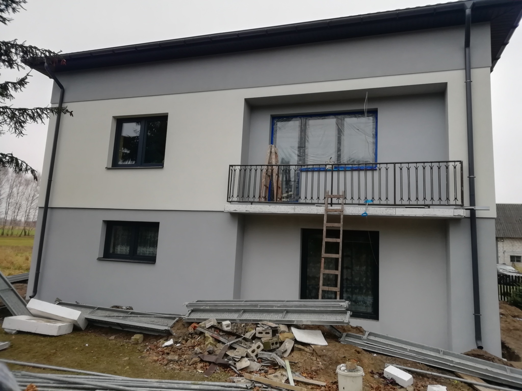 Dwukondygnacyjny dom z balkonem w trakcie prac elewacyjnych; okna zabezpieczone folią, widoczne rusztowania, styropian i gruz na ziemi.