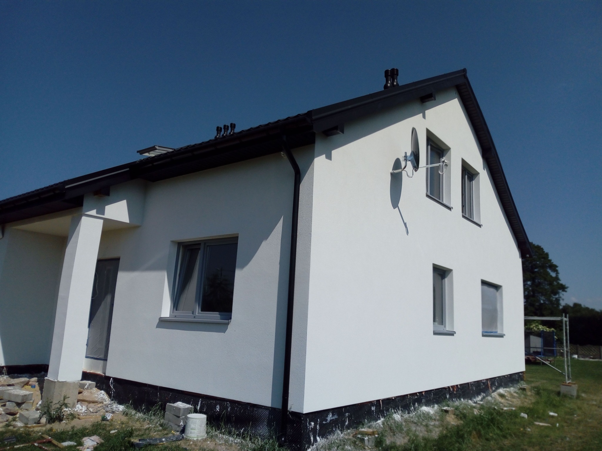 Otynkowana, biała elewacja domu jednorodzinnego z czarnymi elementami wykończeniowymi, widoczne okna z szarymi ramami, fragment dachu i anteny satelitarne, teren po pracach wykończeniowych.