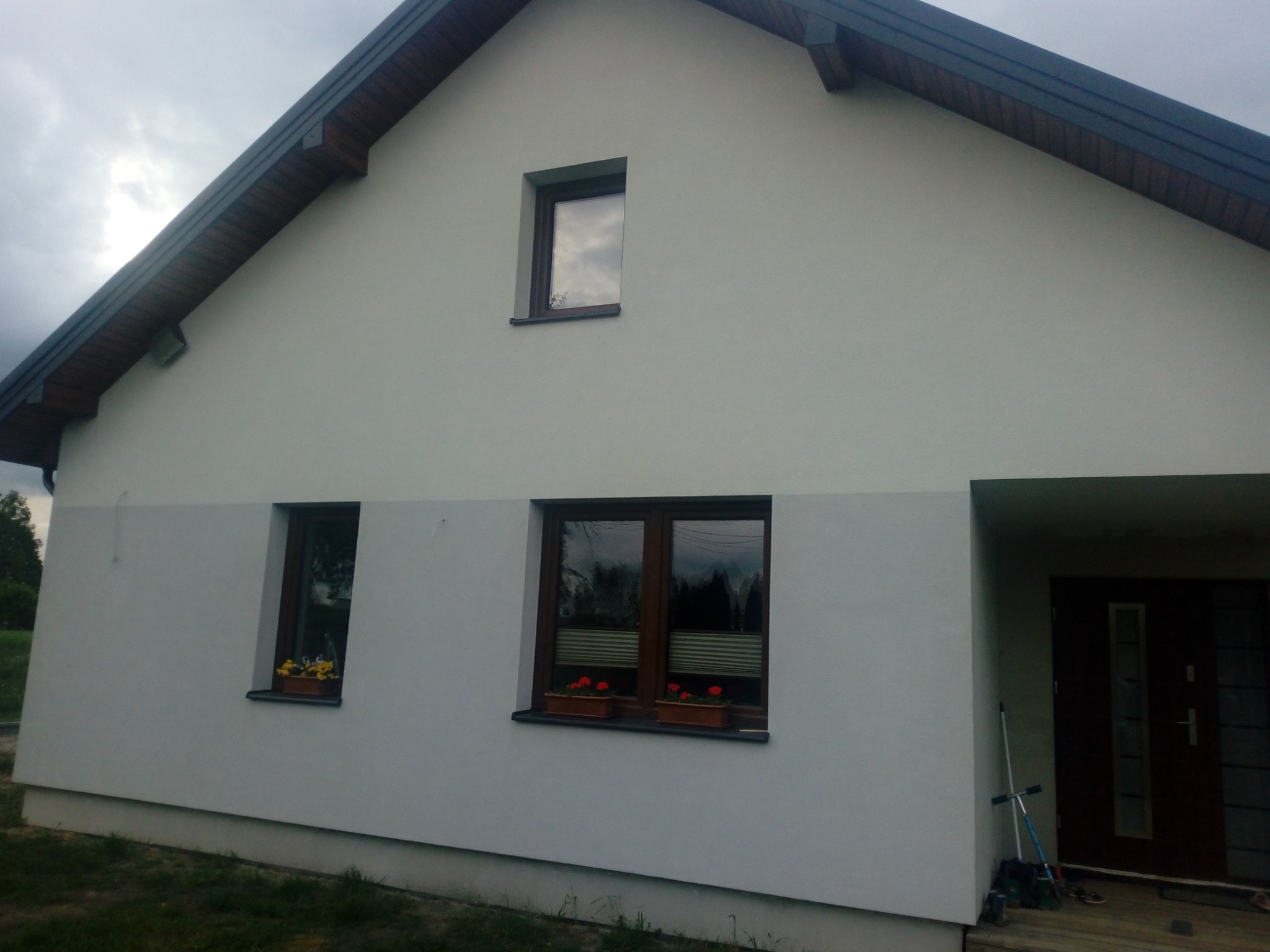 Ocieplony dom jednorodzinny z szarą elewacją, brązową stolarką okienną i kwiatami w donicach pod oknami, widok z boku.