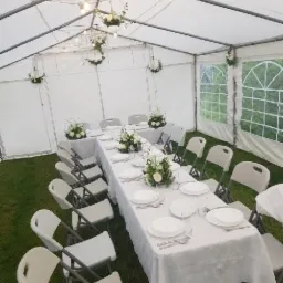 Udekorowany stół z białym obrusem i zastawą, otoczony krzesłami w namiocie z oświetleniem i kwiatowymi dekoracjami.