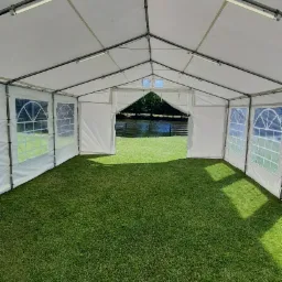 Wnętrze białego namiotu eventowego z oknami i sztuczną trawą, widok na zewnątrz na zieleń i wodę.