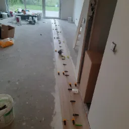 Wyrównywanie podłogi samopoziomującą wylewką z użyciem klipsów poziomujących, widoczne narzędzia i materiały budowlane w surowym wnętrzu.