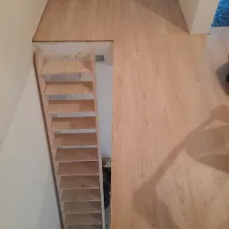 Wykończona podłoga z panelami imitującymi drewno, z widoczną konstrukcją schodów bez wykończenia, prowadzących do niższego poziomu.