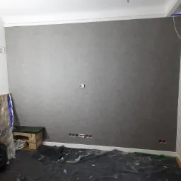 Ściana po tapetowaniu szarą tapetą o delikatnej strukturze, widoczne puszki elektryczne, podłoga zabezpieczona folią malarską, narożnik ściany z pionowym pasem tapety w innym wzorze.