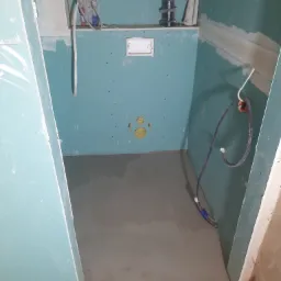 Niedokończona łazienka z zielonymi płytami gipsowo-kartonowymi na ścianach, widoczne instalacje wodne i kanalizacyjne, świeżo wylana wylewka na podłodze, widoczne łączenia płyt wymagające wykończenia.