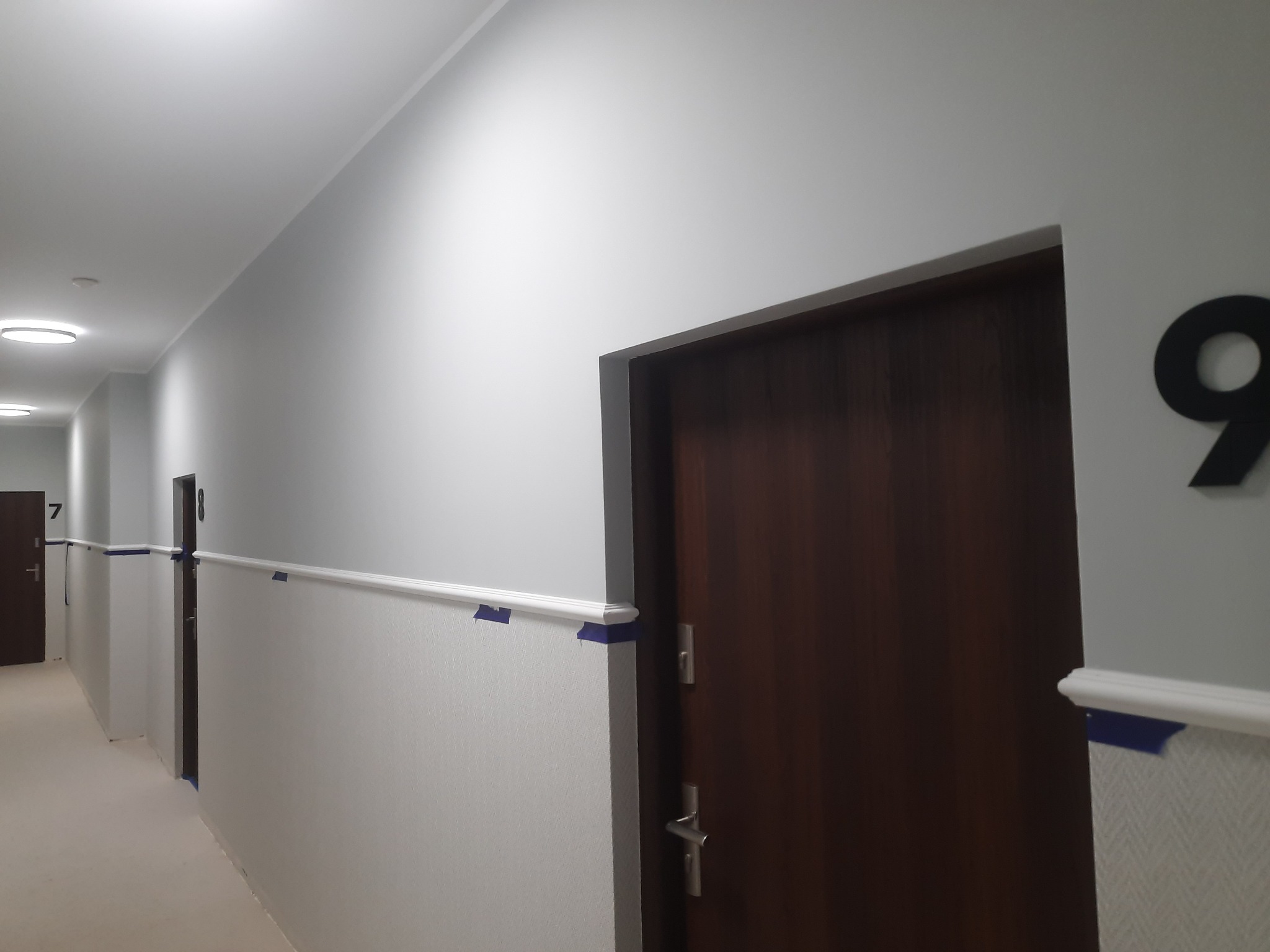Korytarz w trakcie malowania, z zabezpieczonymi listwami przypodłogowymi niebieską taśmą malarską i świeżo pomalowanymi szarymi ścianami, widoczne drzwi do pokoi z numerami.