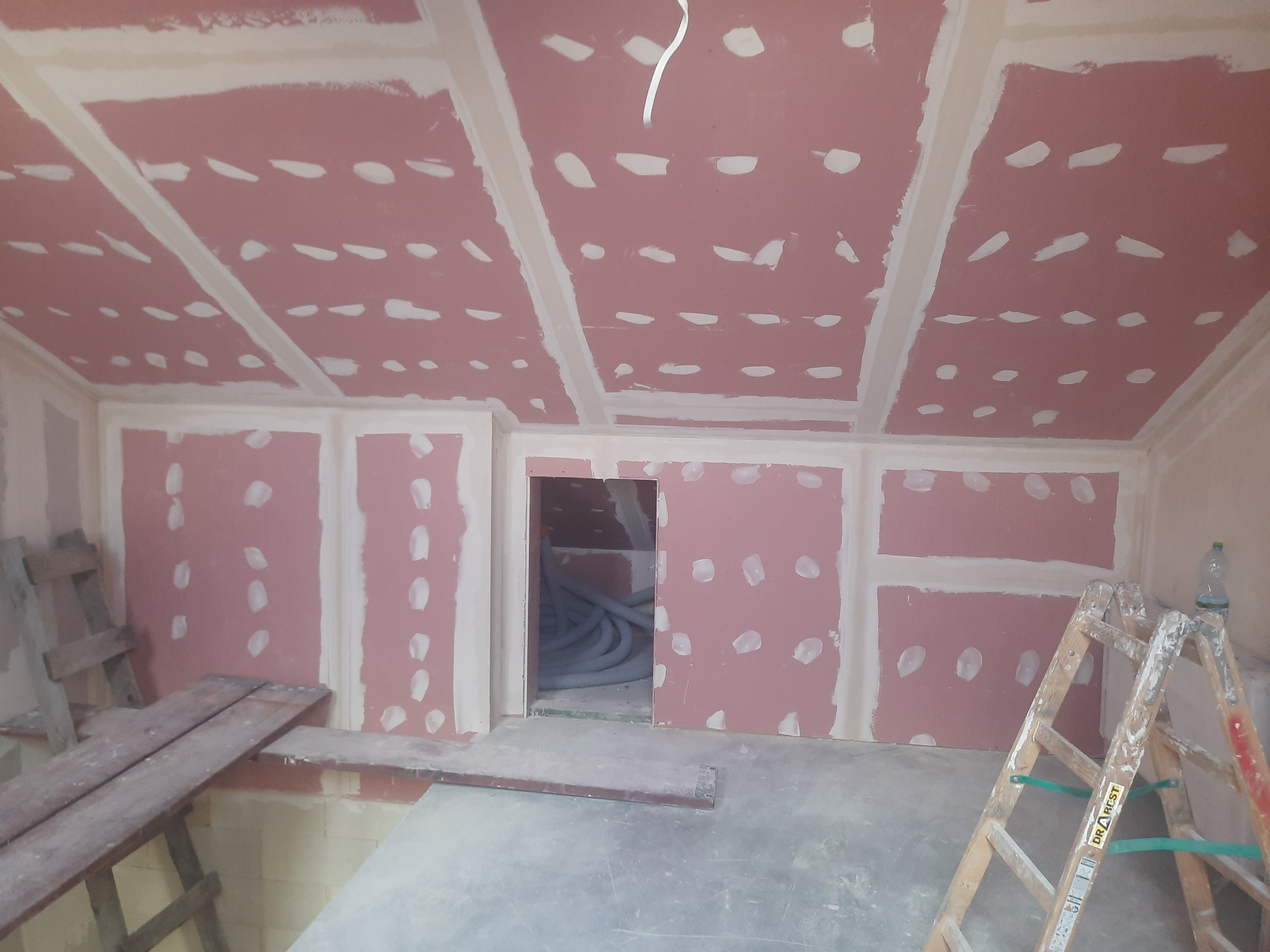 Poddasze w trakcie wykańczania: zamontowane płyty z widocznymi miejscami na wkręty, drewniany pomost roboczy i drabina przystawna z butelką wody w rogu pomieszczenia.