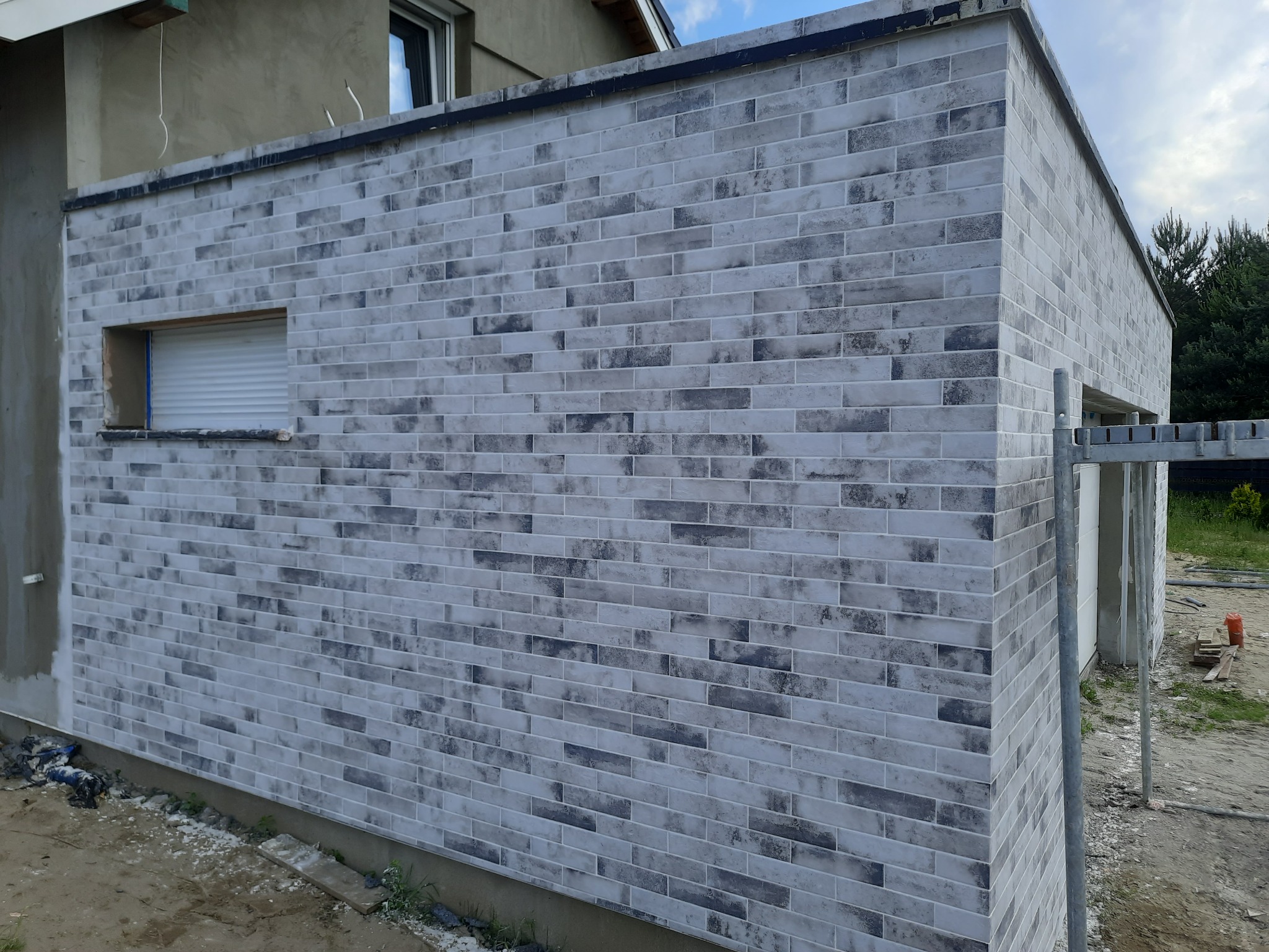 Elewacja budynku wykończona dekoracyjną cegłą klinkierową w odcieniach szarości, widoczny fragment okna z roletą zewnętrzną oraz rusztowanie.