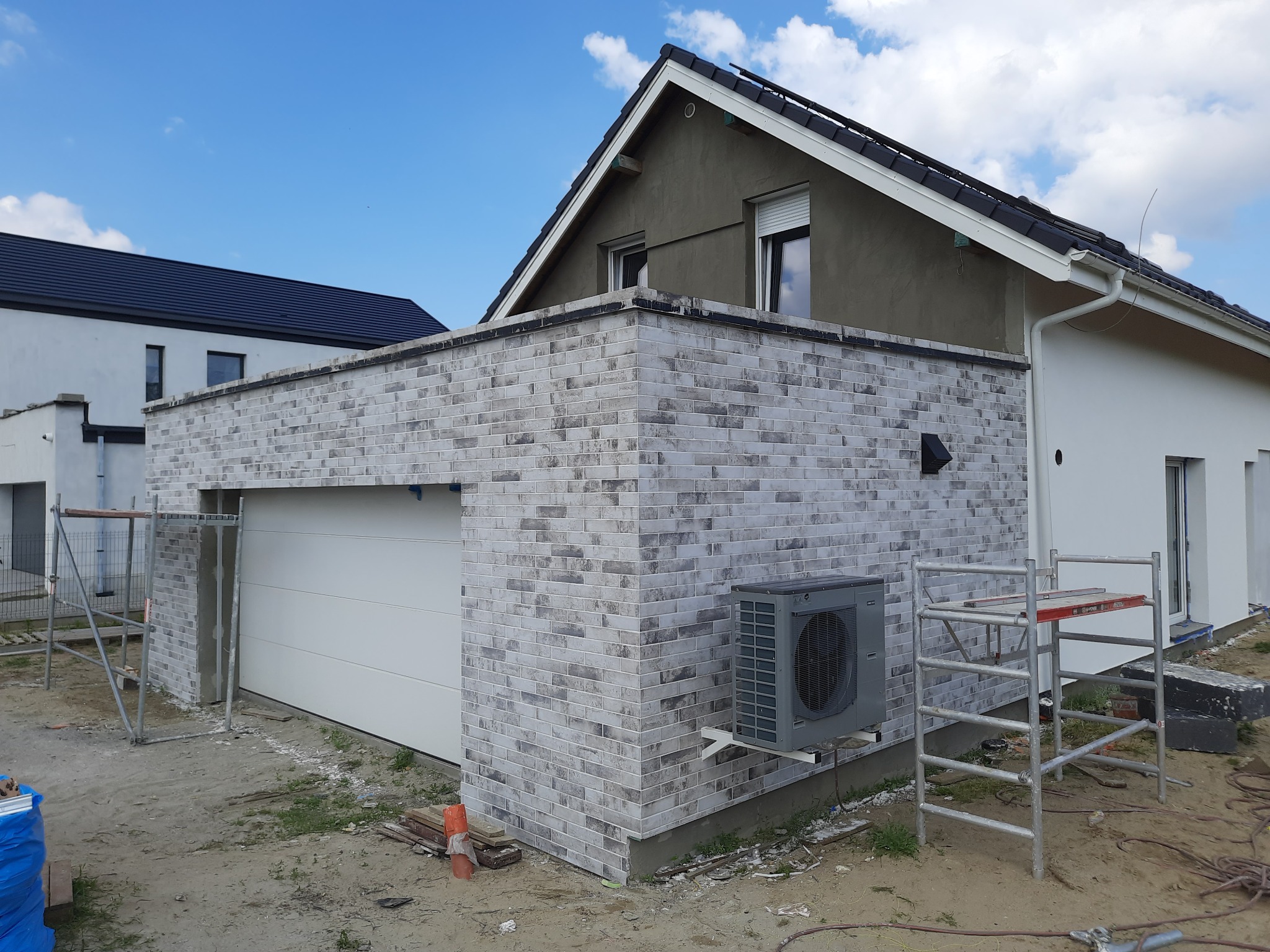 Budynek z garażem w trakcie wykończenia elewacji z szarej cegły, widoczne rusztowania i klimatyzator zewnętrzny.