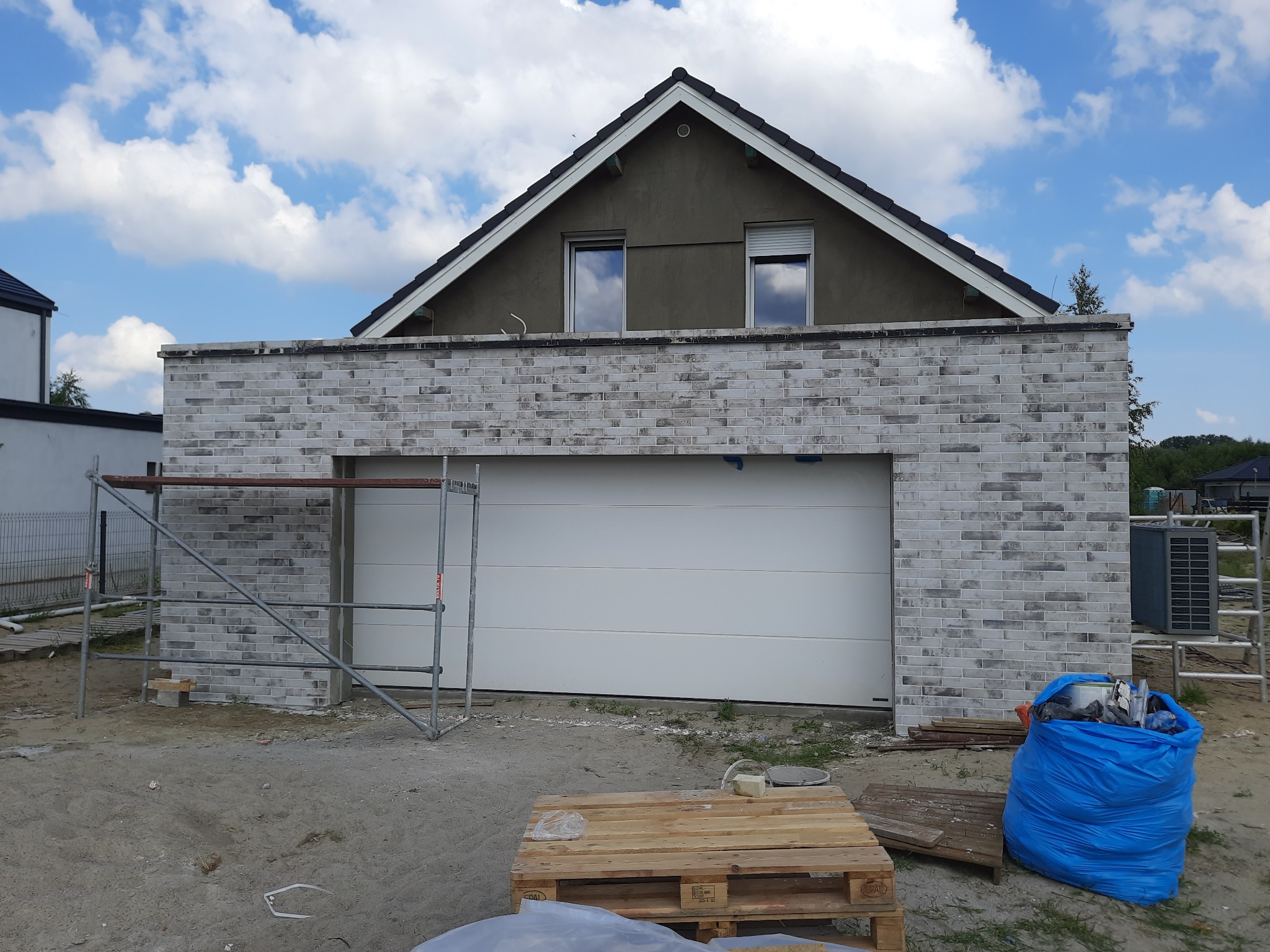 Niedokończony garaż z białej cegły, rusztowanie z lewej strony, paleta i niebieski worek z odpadami budowlanymi na pierwszym planie, poddasze domu w tle, błękitne niebo z chmurami.