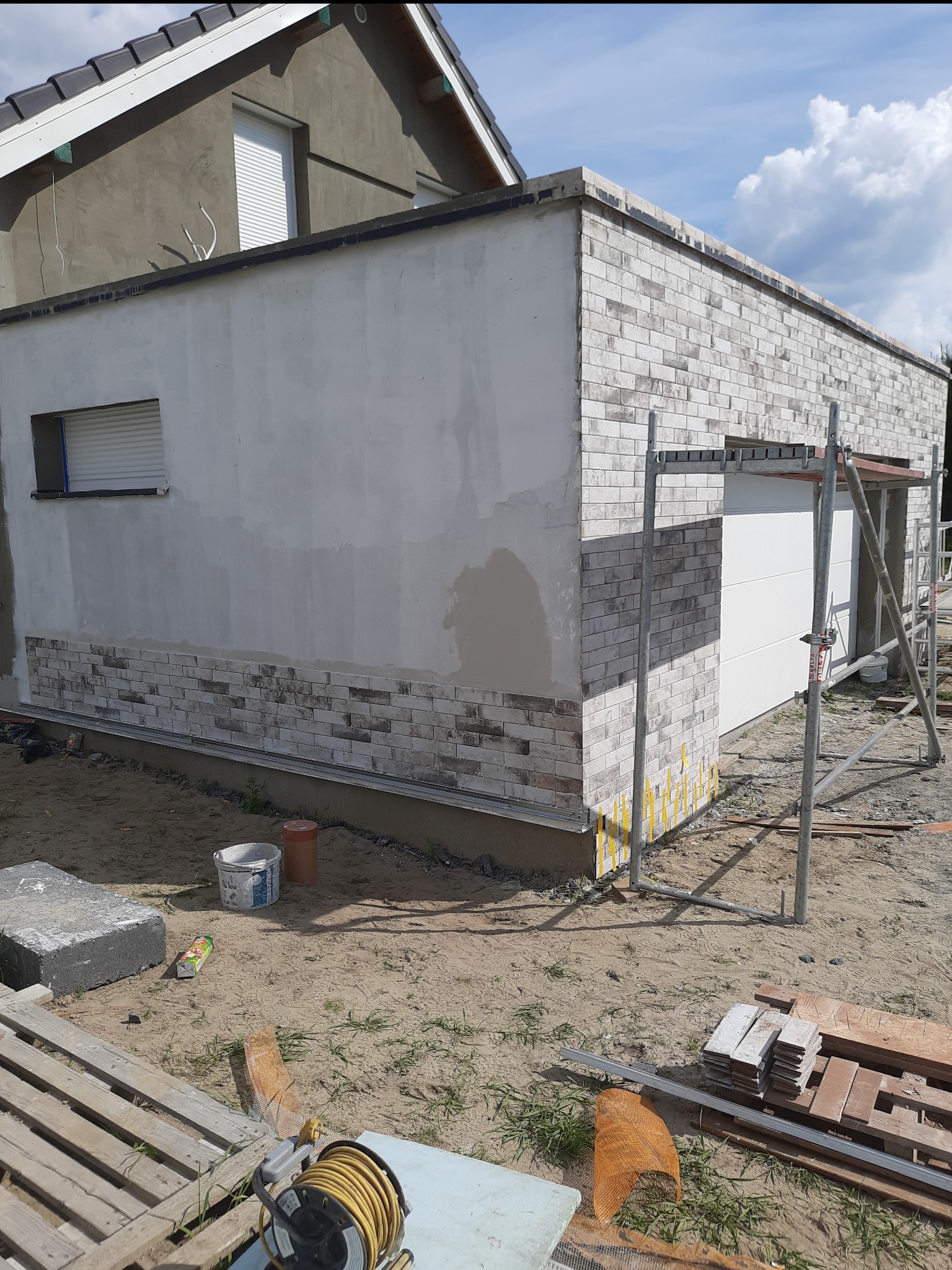 Budynek gospodarczy w trakcie wykończenia elewacji: częściowo otynkowana ściana, fragment obłożony dekoracyjną cegłą, widoczne rusztowanie i materiały budowlane.