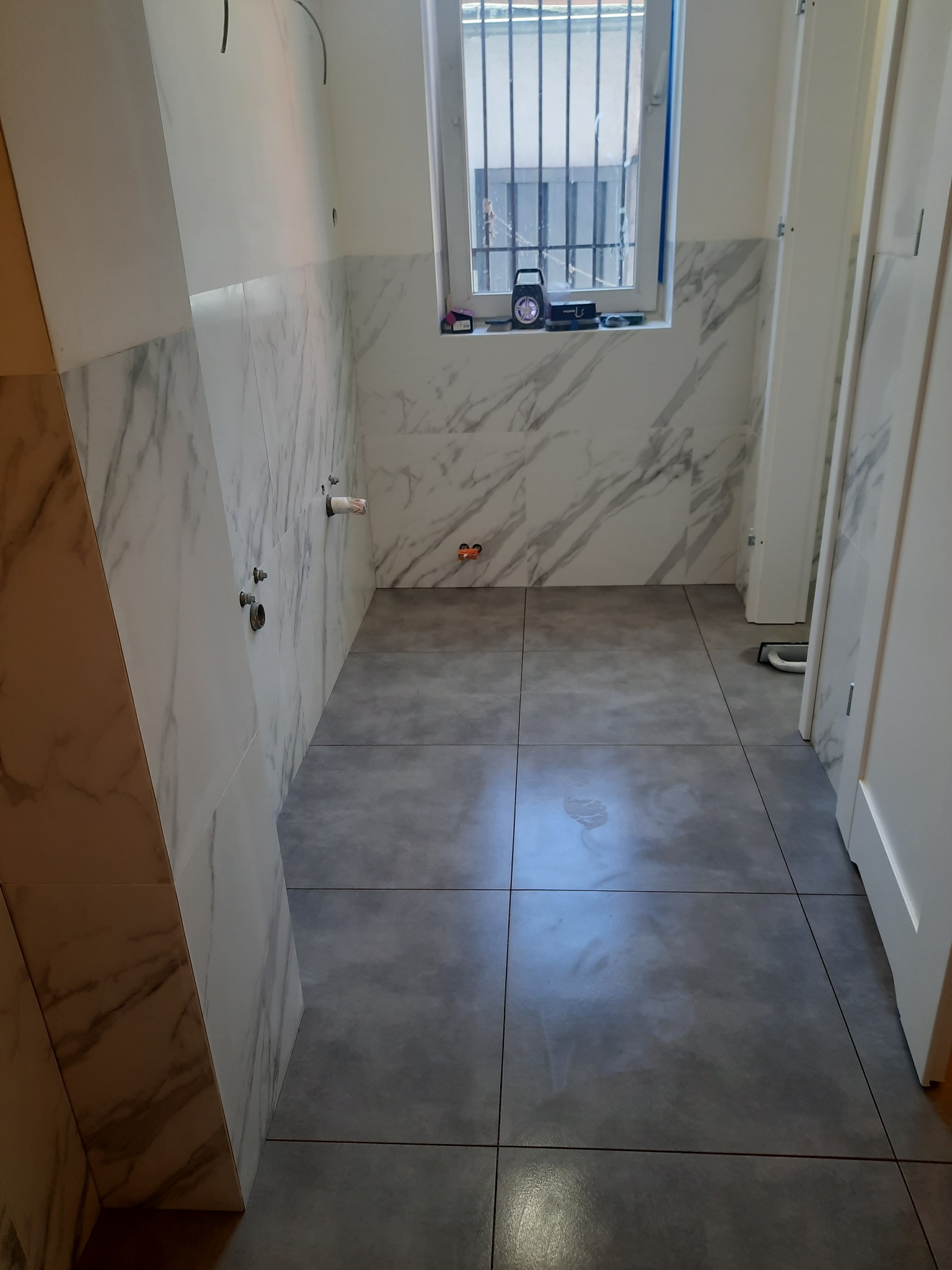 Wykończona łazienka z szarymi płytkami podłogowymi i białymi płytkami imitującymi marmur na ścianach, widoczne wystające rury i otwory na baterię, na parapecie stoi głośnik przenośny.