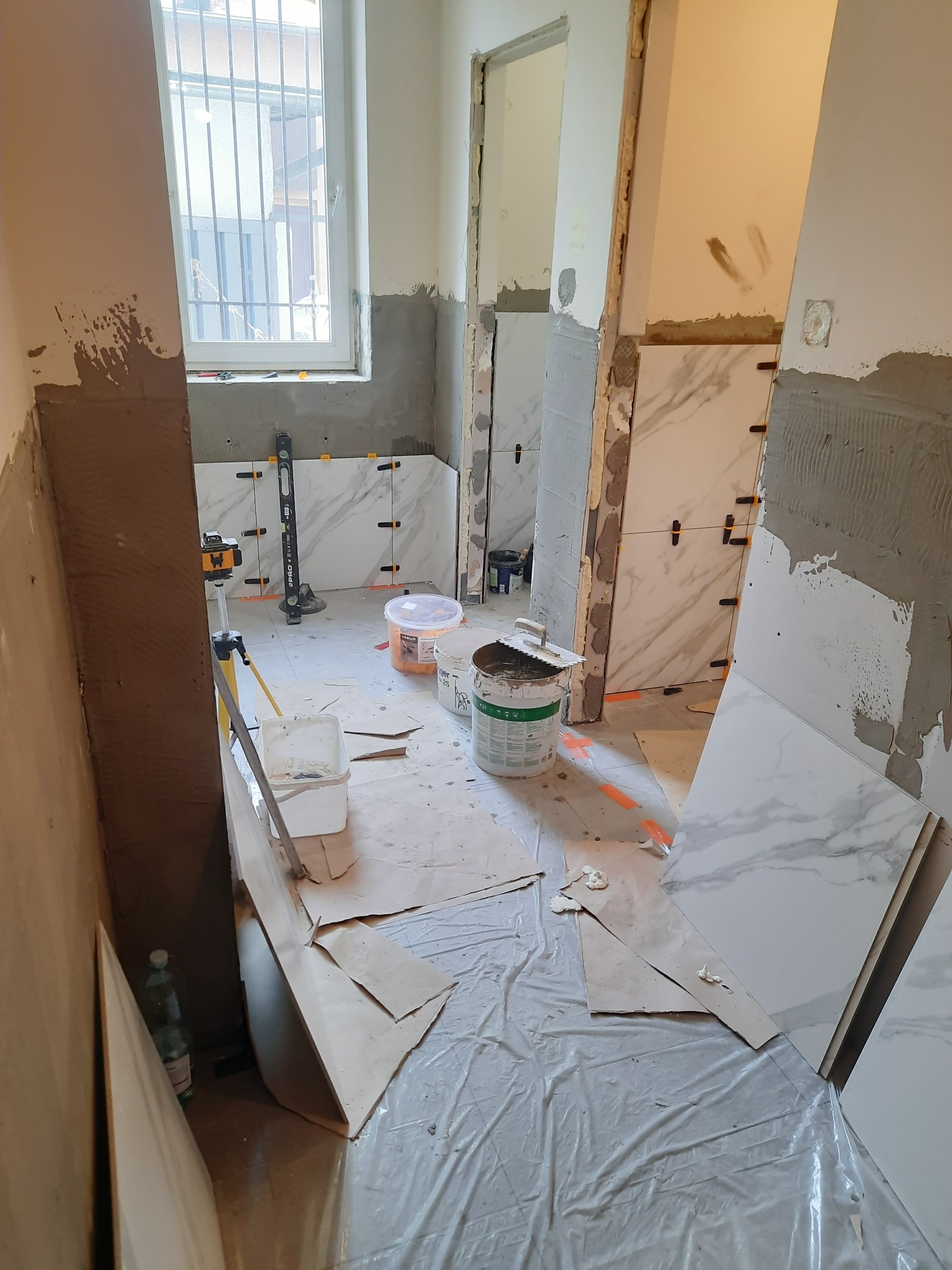 Remont łazienki w trakcie: białe płytki imitujące marmur układane na ścianach, widoczne narzędzia glazurnicze, poziomica laserowa, wiaderka z zaprawą, folia ochronna na podłodze.