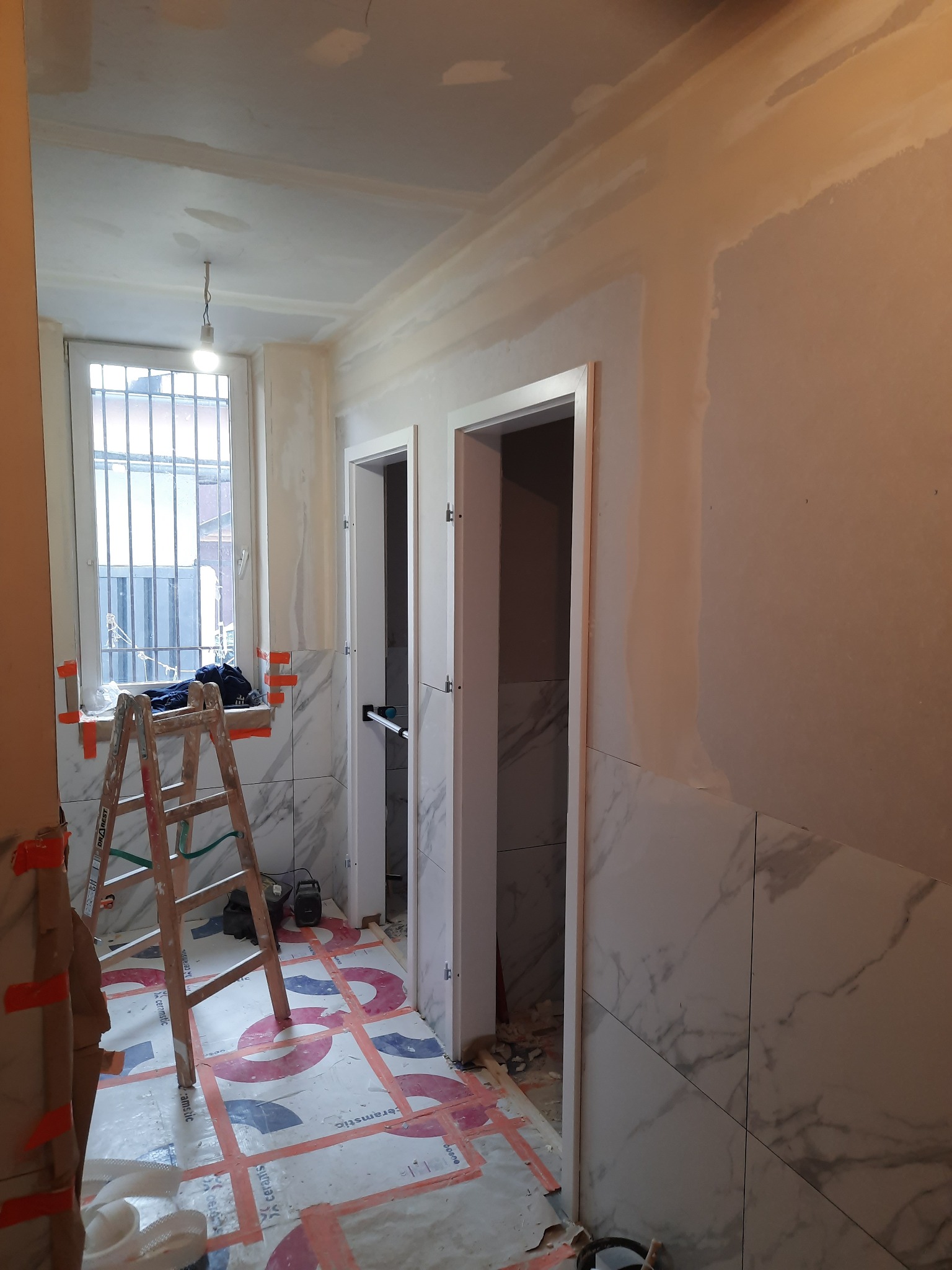 Wnętrze łazienki w trakcie remontu: widoczne ślady prac na ścianach i suficie, częściowo położone płytki imitujące marmur, zabezpieczona podłoga, drabina oparta o okno z kratą.