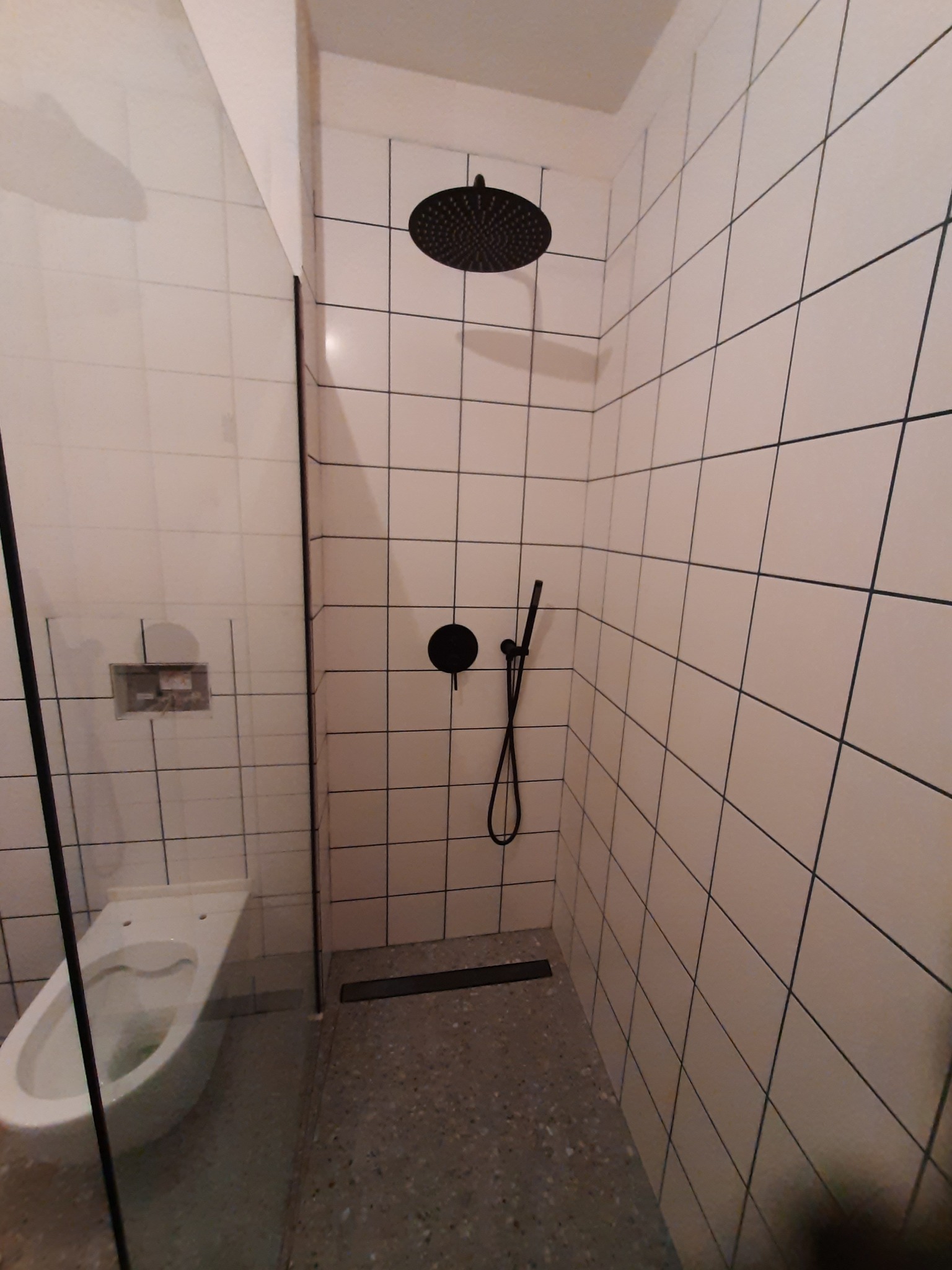 Nowoczesna kabina prysznicowa z czarną armaturą i białymi płytkami, widoczny fragment toalety za szklaną ścianką.