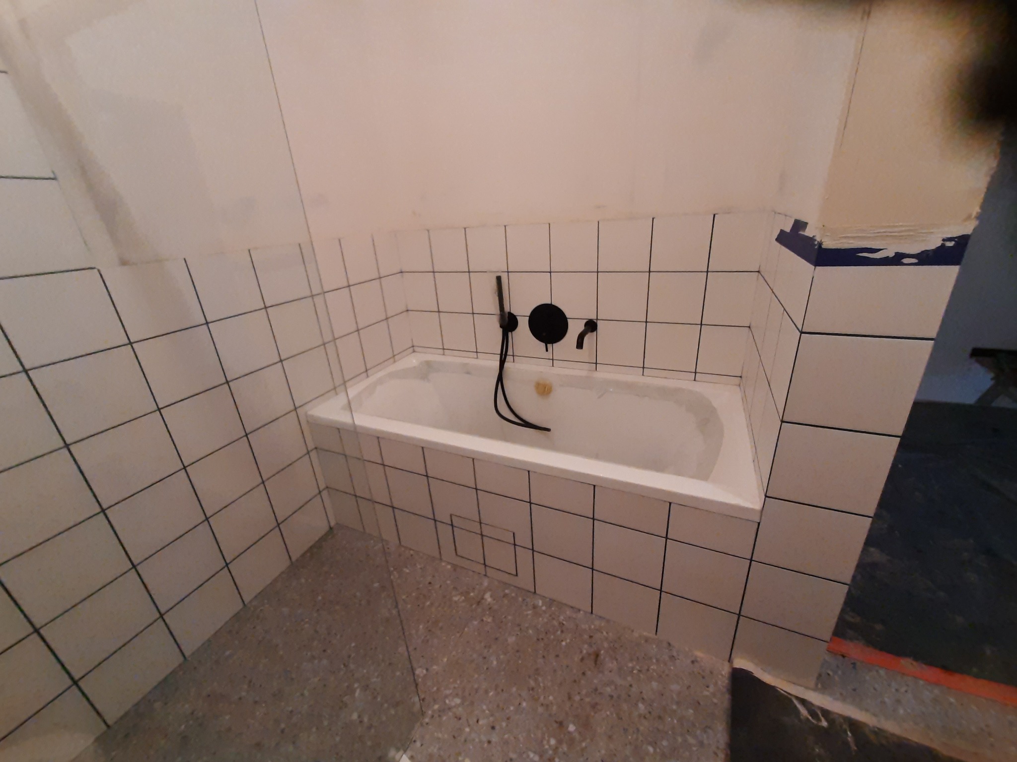 Wykończona łazienka z białą wanną obudowaną płytkami w kratkę, czarna armatura, surowe ściany w trakcie przygotowania do malowania i terakota na podłodze.