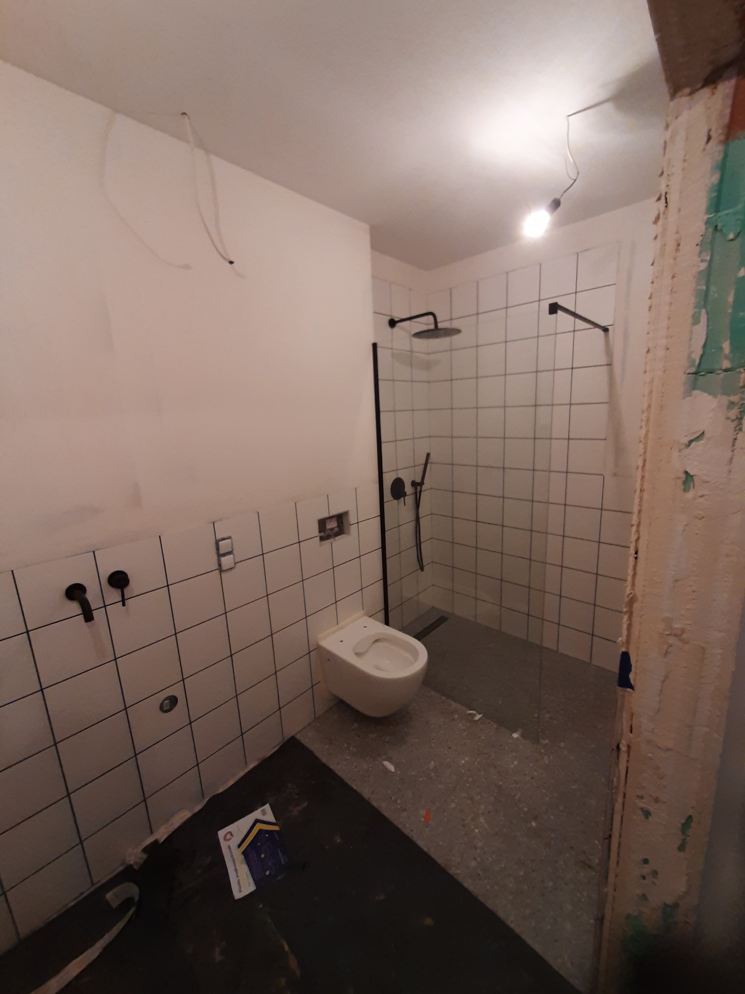 Remont łazienki: widok na kabinę prysznicową typu walk-in z czarną armaturą, wiszącą toaletę i surowe ściany z resztkami starej farby