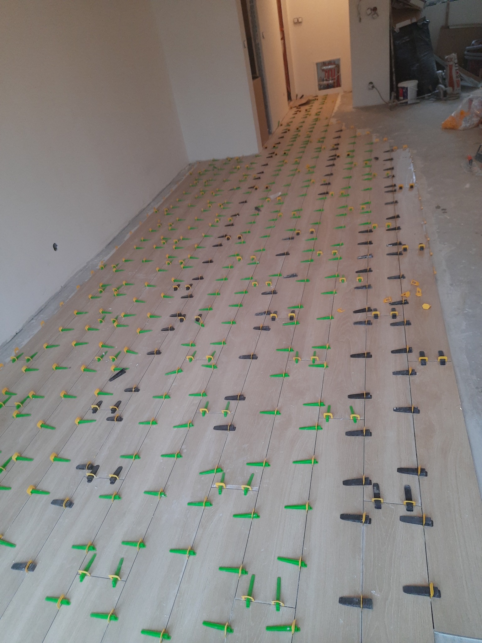 Układanie podłogi z płytek ceramicznych z użyciem systemu poziomowania płytek, widoczne kliny i klipsy dystansowe, perspektywa od ściany w głąb pomieszczenia.