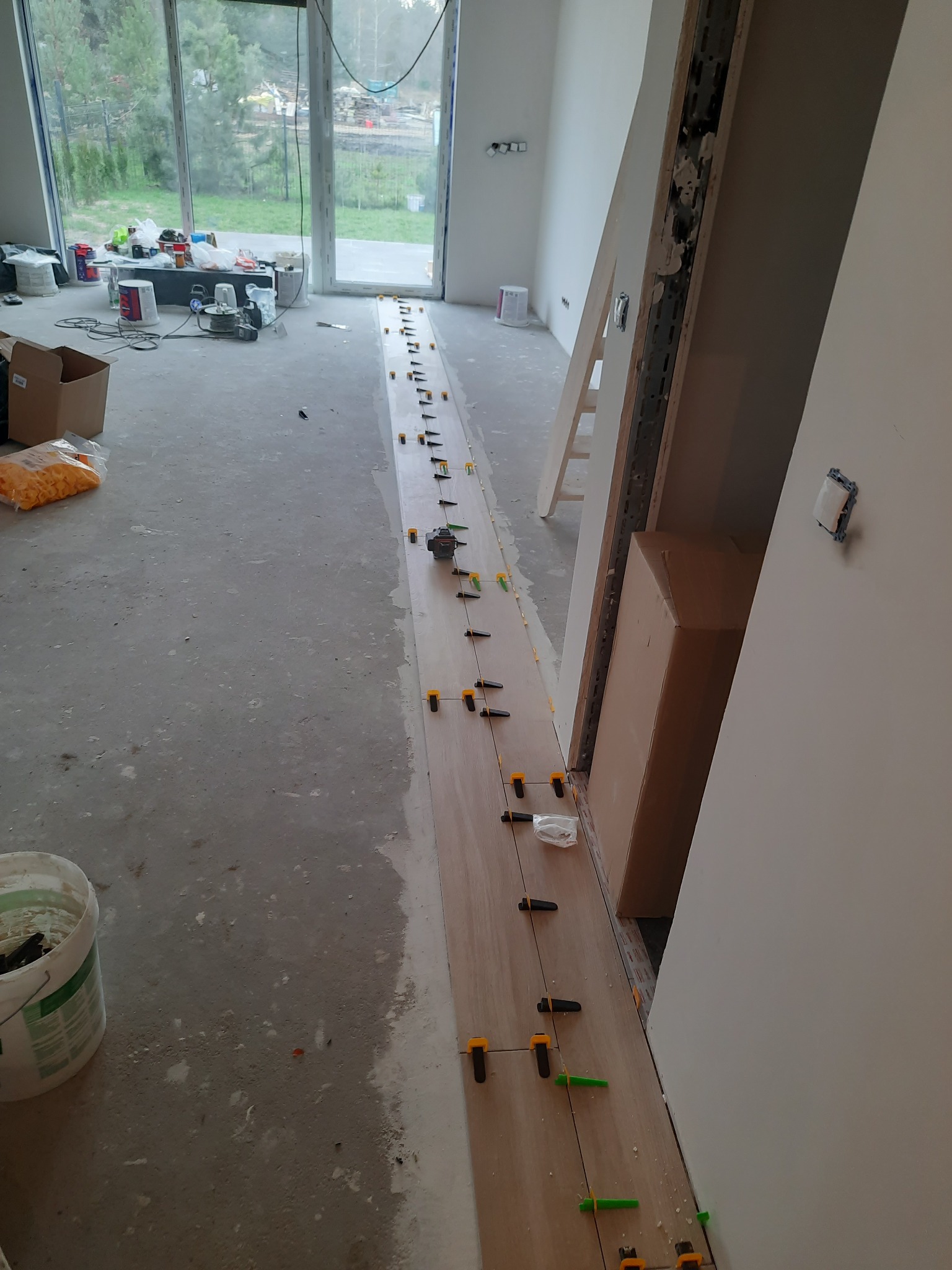 Wyrównywanie podłogi samopoziomującą wylewką z użyciem klipsów poziomujących, widoczne narzędzia i materiały budowlane w surowym wnętrzu.