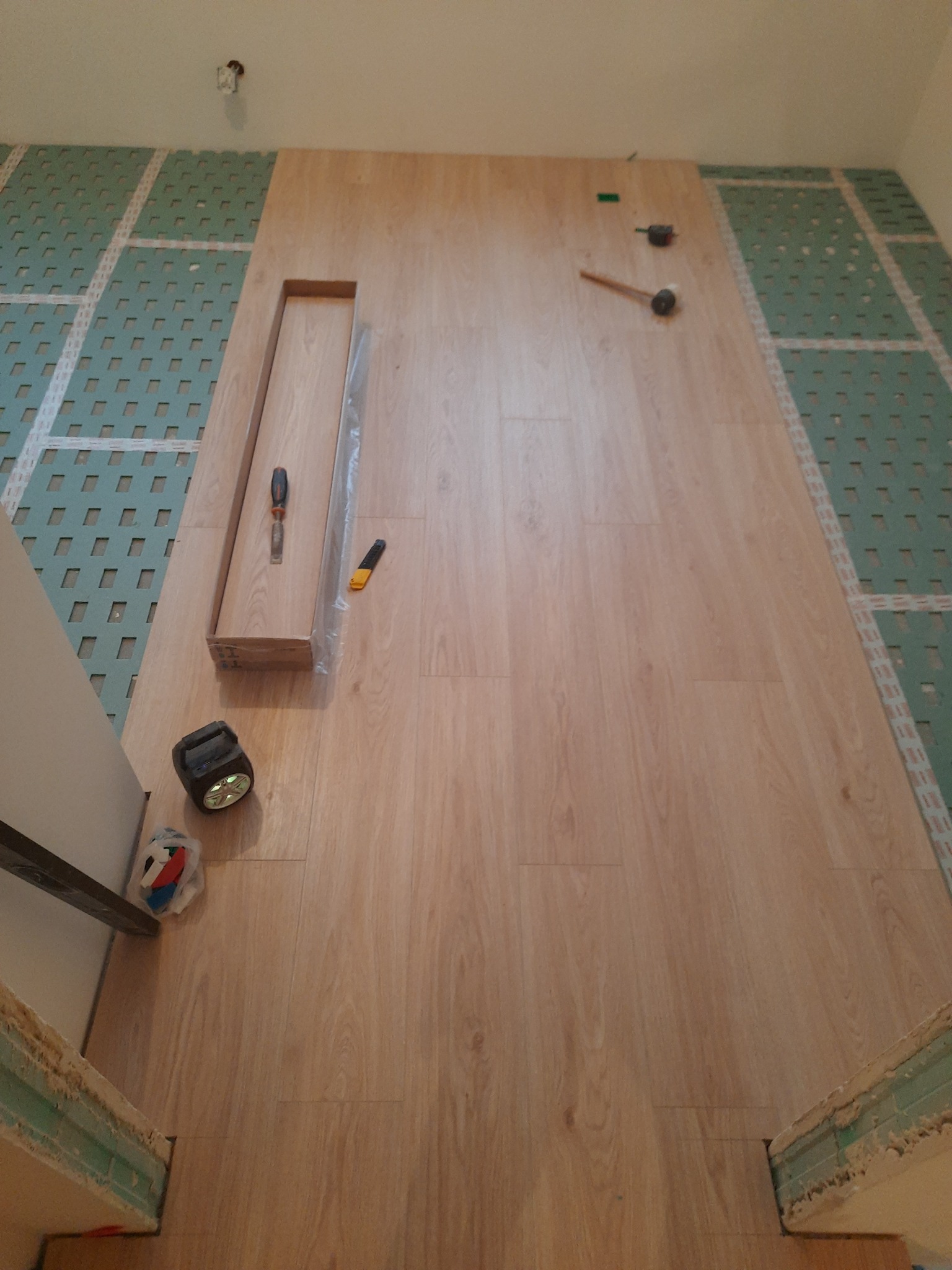 Montaż paneli podłogowych: widok z góry na częściowo ułożone panele o jasnym odcieniu drewna, podkład z zielonymi kwadratami, narzędzia (młotek gumowy, śrubokręt, nóż) leżące na panelach, pudełko...