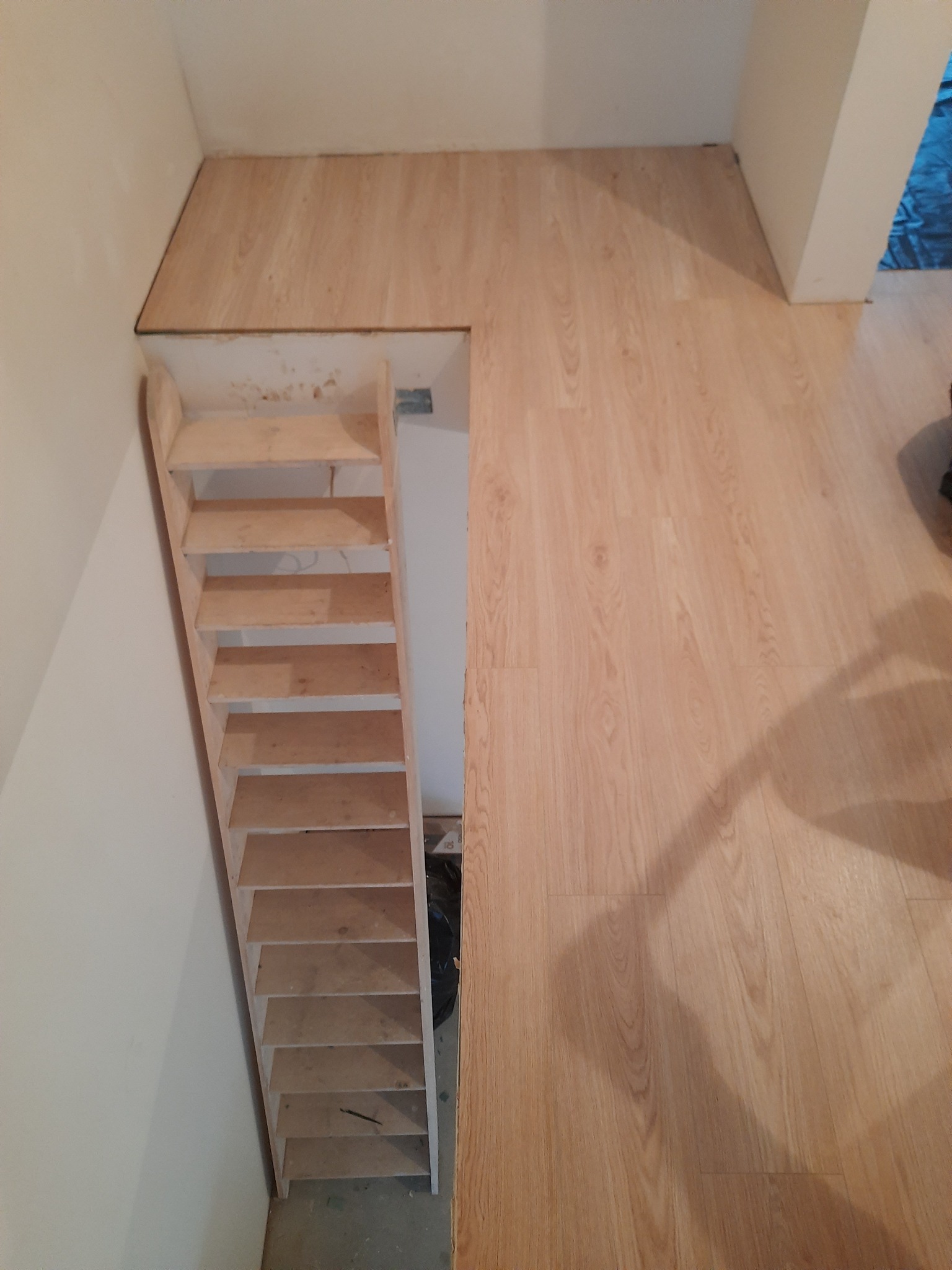 Wykończona podłoga z panelami imitującymi drewno, z widoczną konstrukcją schodów bez wykończenia, prowadzących do niższego poziomu.