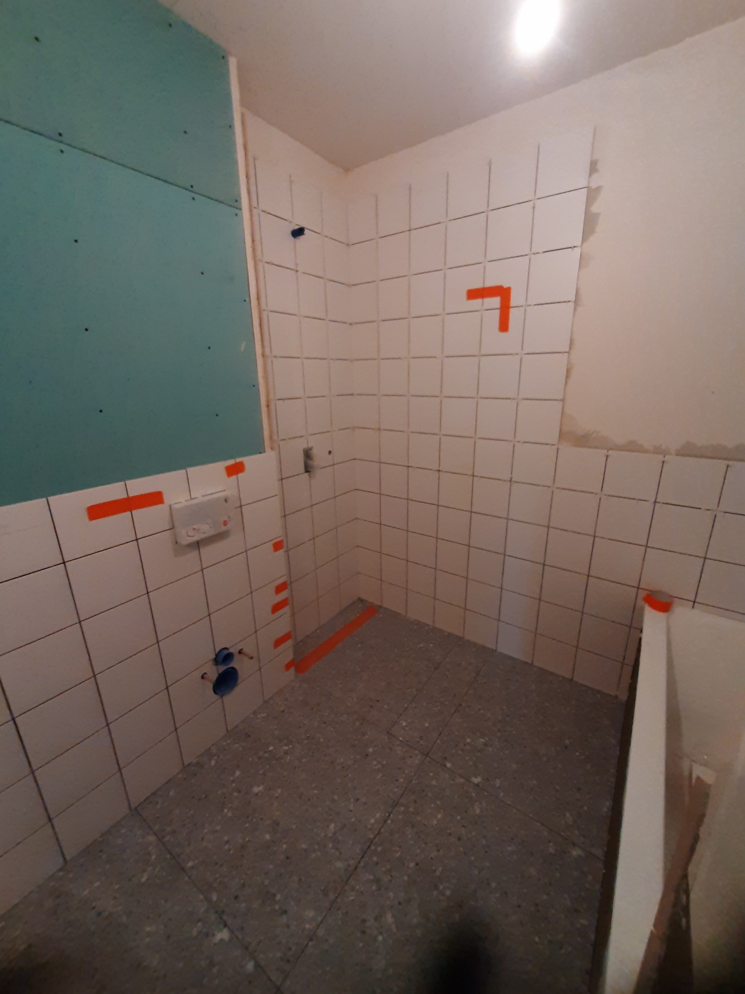 Remont łazienki w trakcie: fragment ściany z zieloną płytą, białe płytki z pomarańczową taśmą wyznaczającą poziom, szara podłoga z lastryko, widoczne rury instalacji wodnej.