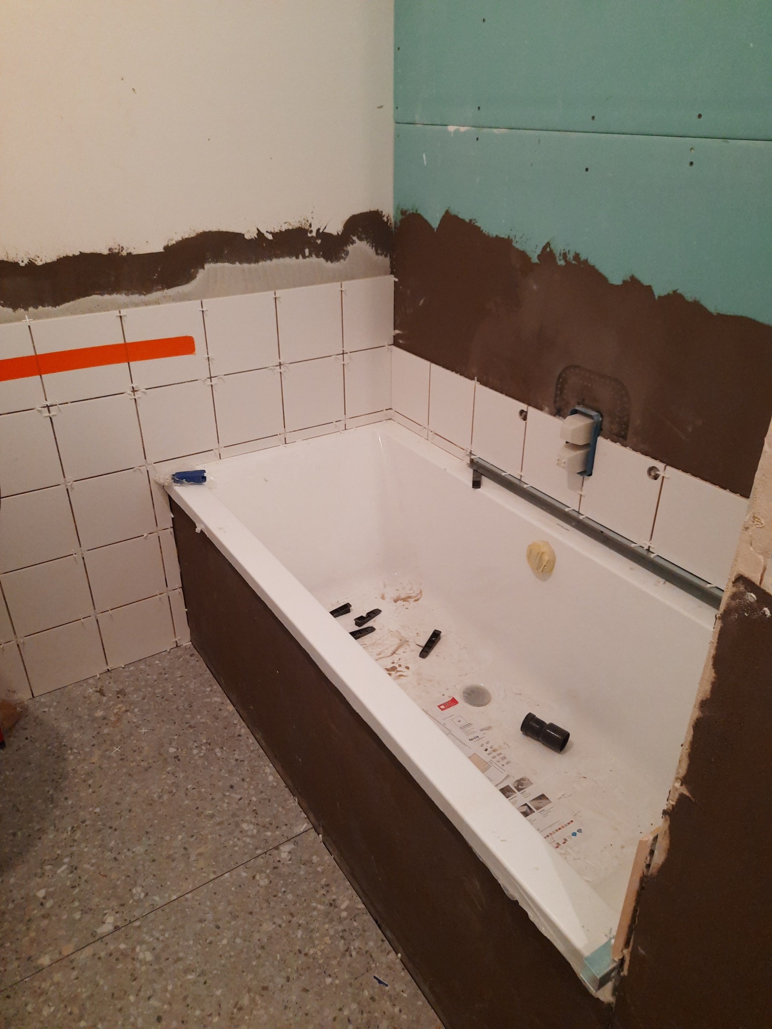 Remont łazienki w trakcie: widoczna wanna, częściowo położone płytki z pomarańczową taśmą, fragment płyty kartonowo-gipsowej z nałożoną warstwą brązowej masy, elementy instalacji w wannie, podłoga...