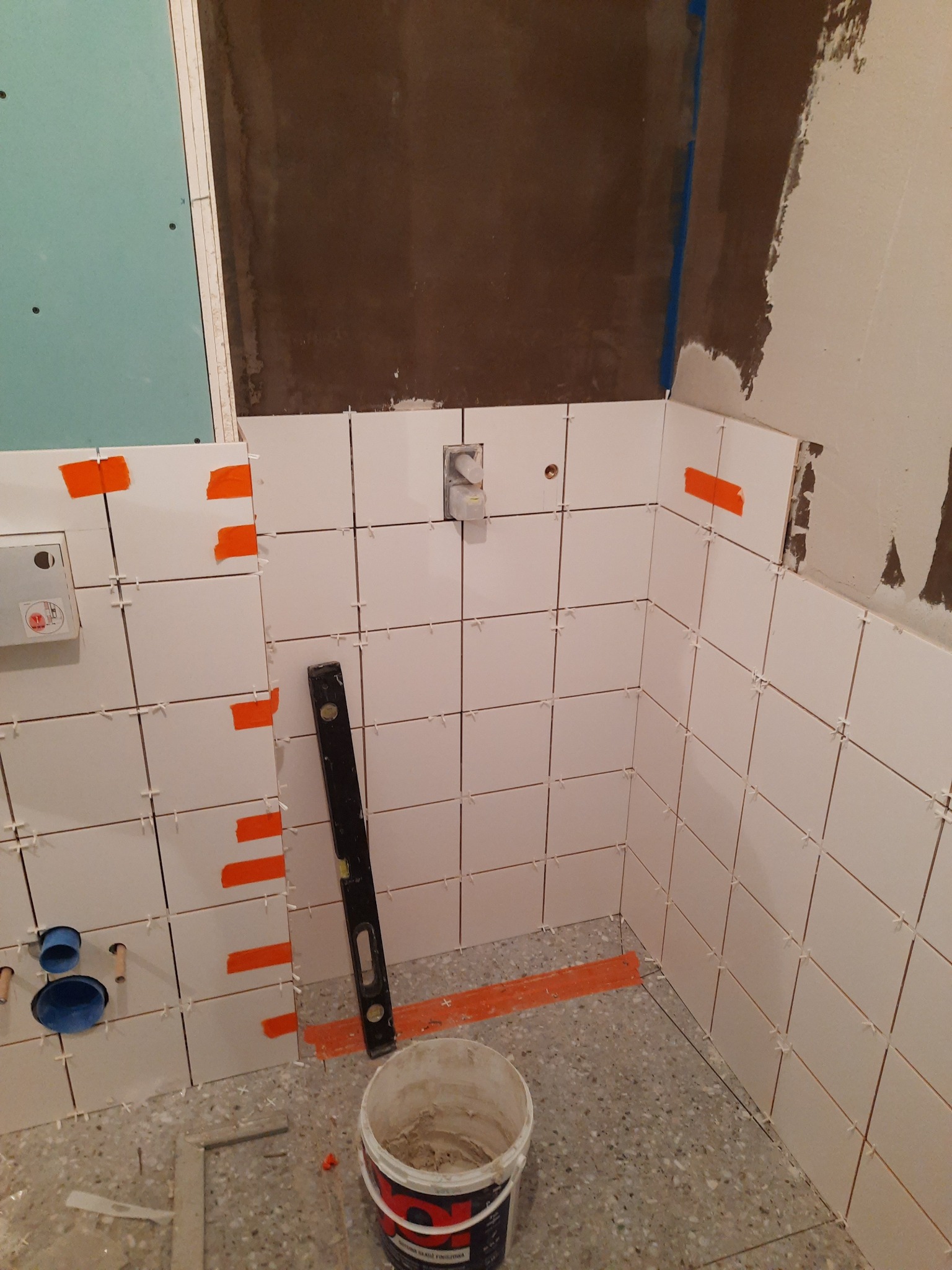 Układanie białych płytek w narożniku łazienki, z użyciem pomarańczowej taśmy maskującej, poziomicy i pojemnika z zaprawą klejową. Widoczne krzyżyki dystansowe i otwory na instalacje.