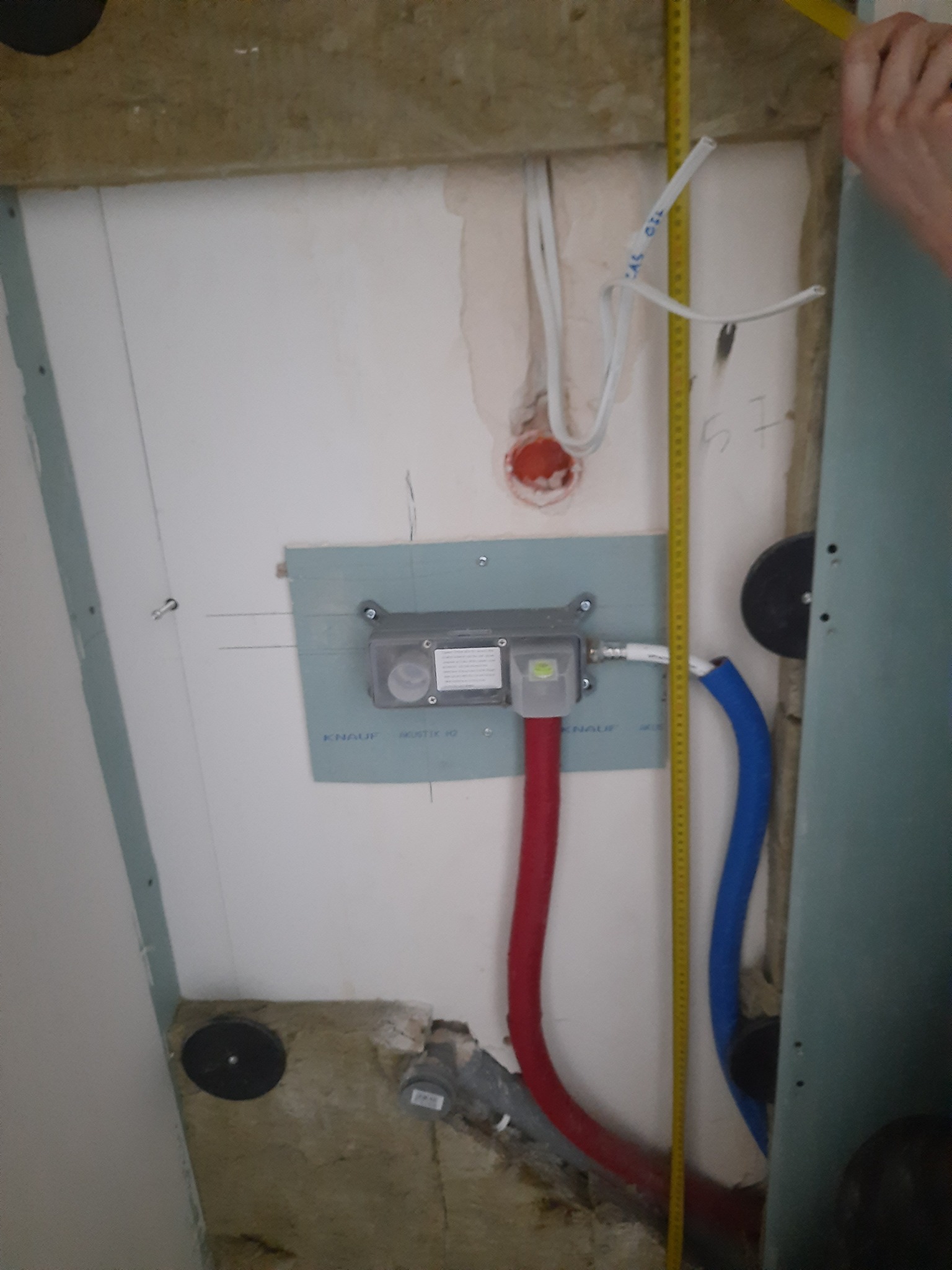 Instalacja podtynkowa z widocznymi przewodami elektrycznymi, rurami wodnymi (czerwona i niebieska) oraz miernikiem poziomu, przy częściowo zamontowanej płycie gipsowo-kartonowej. W tle surowa...