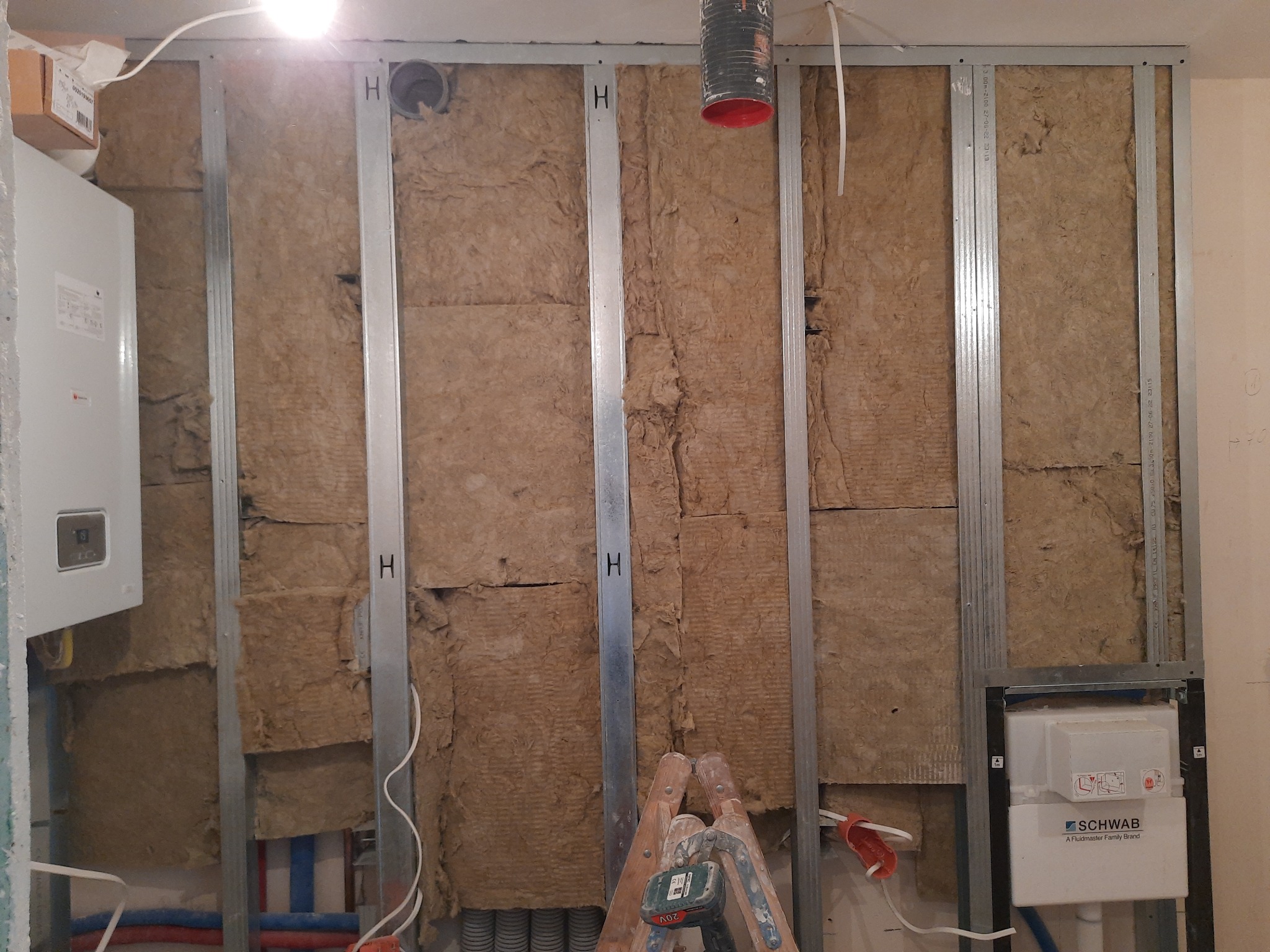 Montaż izolacji z wełny mineralnej w metalowym stelażu, widoczna drabina i instalacja sanitarna, w tle zbiornik WC.