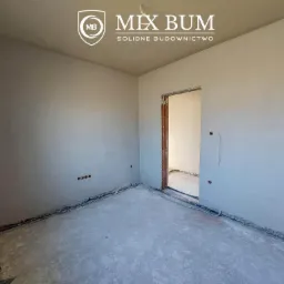 Wnętrze pomieszczenia po nałożeniu tynków maszynowych, widoczne gładkie ściany i surowa podłoga, wystające przewody elektryczne, logo firmy budowlanej w górnej części kadru.