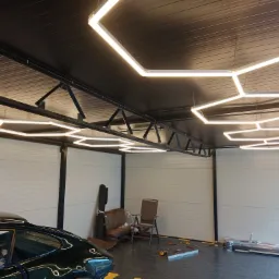 Nowoczesne oświetlenie garażu z heksagonalnymi lampami LED zawieszonymi na łańcuchach, widoczny fragment ciemnozielonego samochodu i wyposażenie warsztatowe.