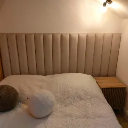 Beżowe, tapicerowane wezgłowie łóżka z pionowymi przeszyciami, widoczne drewniane szafki nocne i dekoracyjne poduszki na białej narzucie w sypialni z drewnianym słupem.