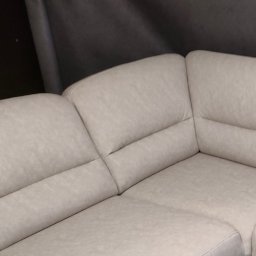 Dobromir Tapicer - Fragment jasnej, narożnej sofy z tapicerką o delikatnej fakturze, widoczny szew na łączeniu segmentów i ciemne tło za meblem.