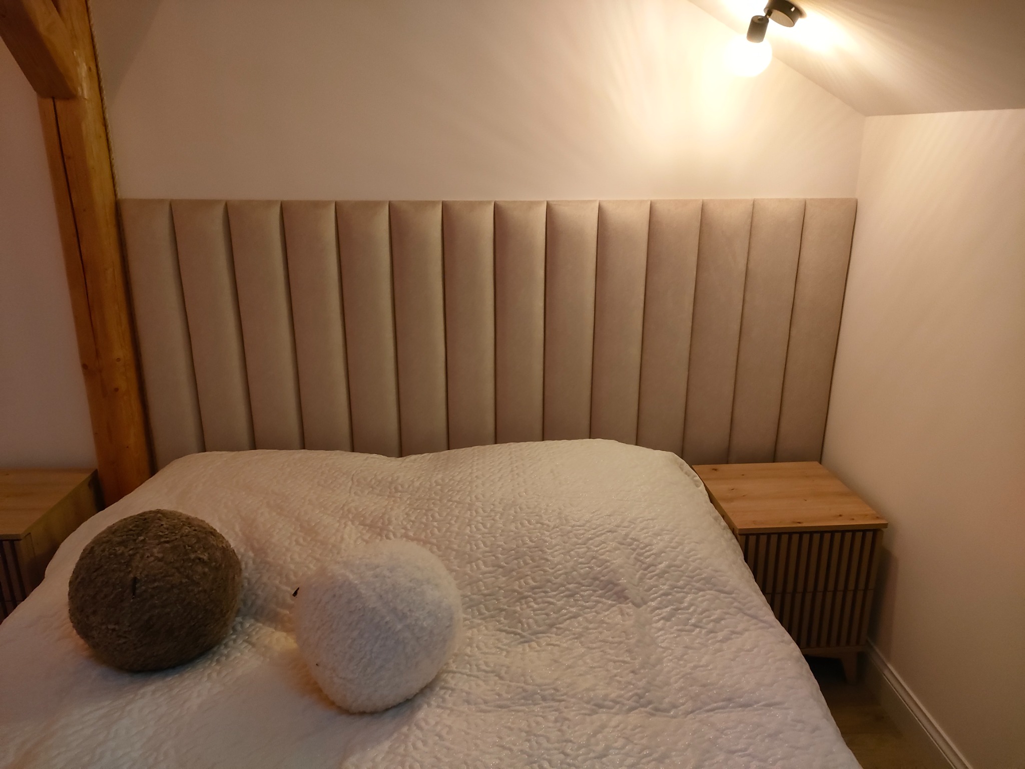 Beżowe, tapicerowane wezgłowie łóżka z pionowymi przeszyciami, widoczne drewniane szafki nocne i dekoracyjne poduszki na białej narzucie w sypialni z drewnianym słupem.