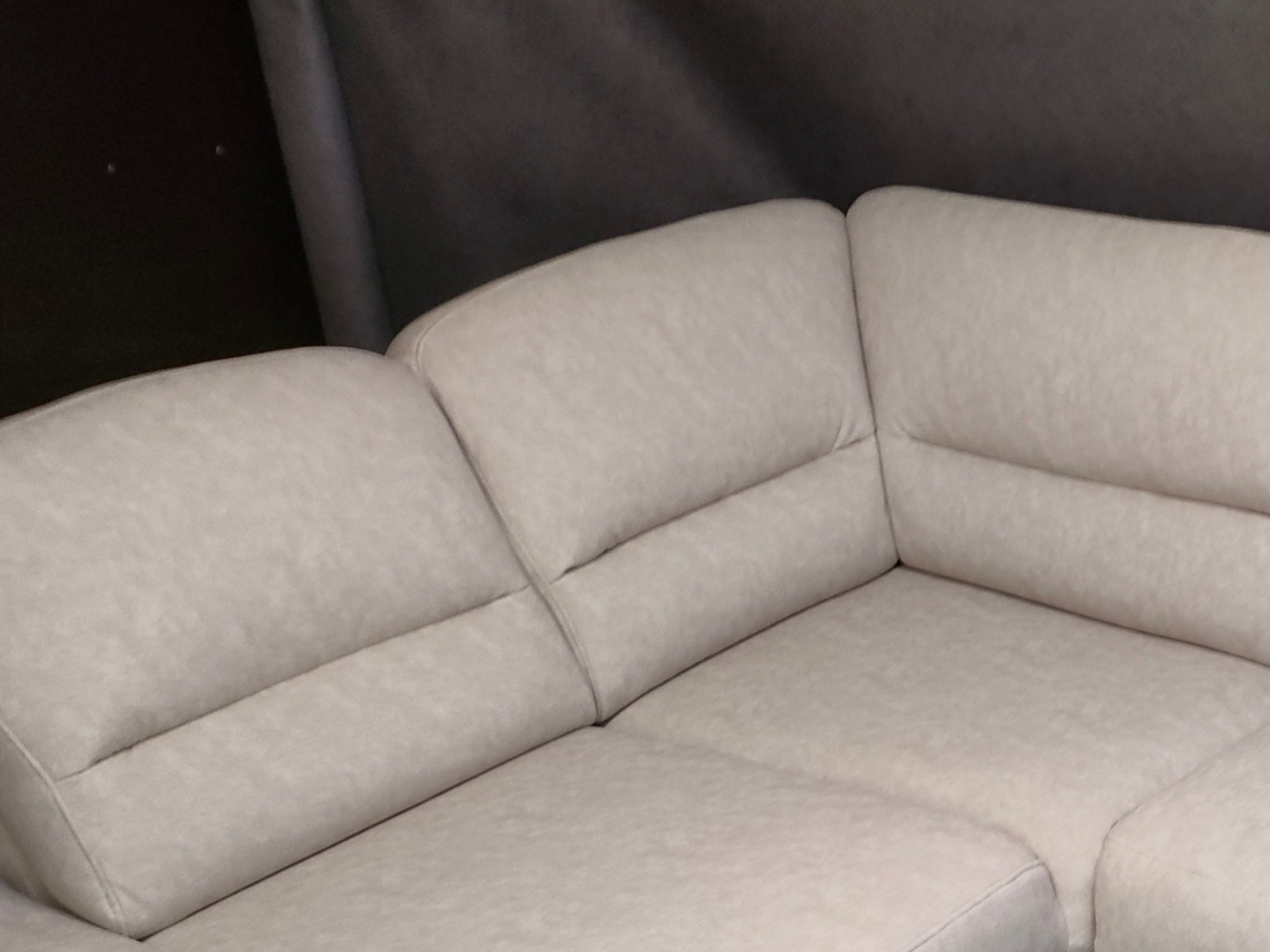 Fragment jasnej, narożnej sofy z tapicerką o delikatnej fakturze, widoczny szew na łączeniu segmentów i ciemne tło za meblem.