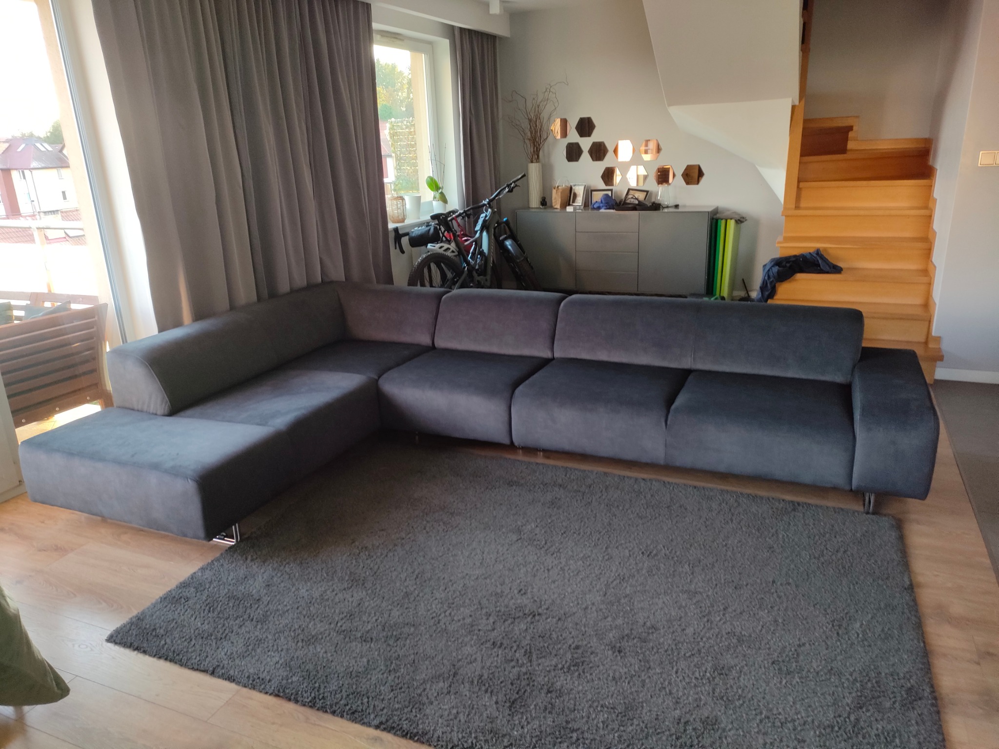 Szary, narożny zestaw wypoczynkowy z niskim oparciem i metalowymi nóżkami w salonie w Żukowie, widok na dywan, schody i rower w tle.