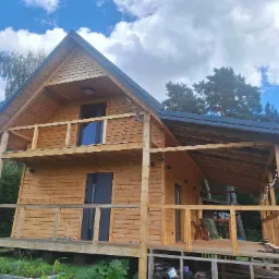 Gotowy, dwukondygnacyjny dom z drewna z balkonem, widoczny z zewnątrz, na tle błękitnego nieba z chmurami i zieleni drzew.