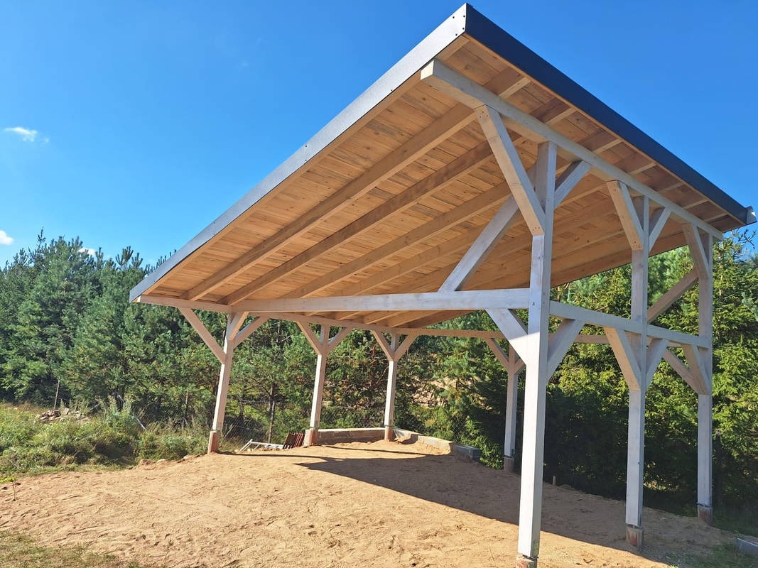 Drewniana konstrukcja z zadaszeniem na piaszczystym podłożu, widok z dołu ukazujący solidne, pomalowane na biało słupy i drewniany dach, w tle zielone drzewa i błękitne niebo.