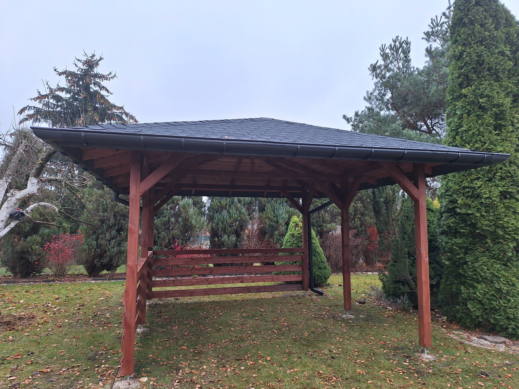 Drewniana konstrukcja z czarnym dachem krytym gontem bitumicznym, otoczona zielenią ogrodu z opadłymi liśćmi na trawie.