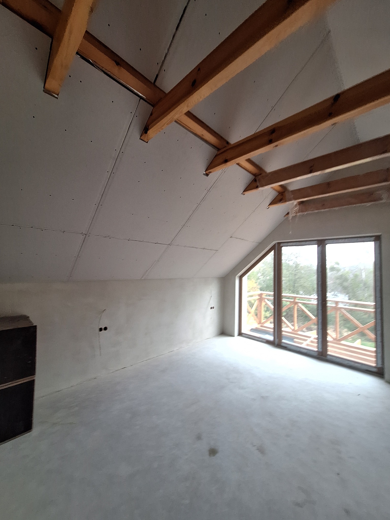 Wnętrze poddasza domu szkieletowego w trakcie budowy: widoczne drewniane belki stropowe, płyty gipsowo-kartonowe na skosach dachu, surowe ściany i okno z widokiem na drewnianą balustradę balkonu.